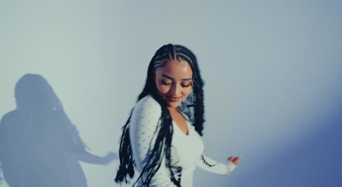 A woman with long braided hair and a white dotted top smiling with eyes closed, standing against a plain wall with her shadow cast on the wall.