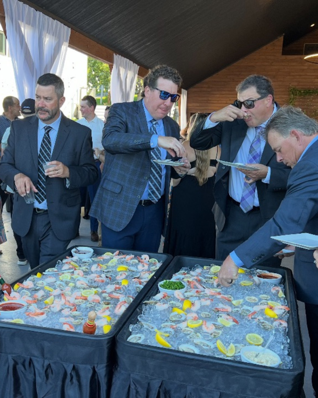 A group of men in suits at a seafood buffet gathering, serving themselves fresh oysters on ice with lemon wedges, at an outdoor event.