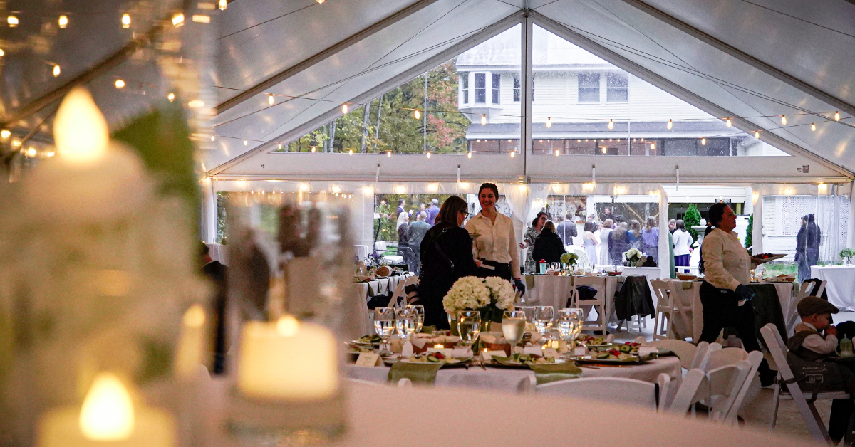 A decorated event tent with tables set for a celebration, featuring floral centerpieces, candles, and string lights, with guests and staff inside and outside.