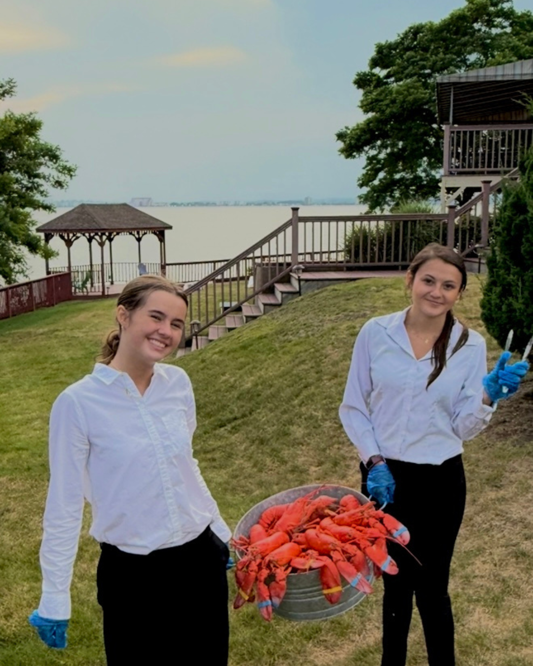 Two women in white shirts and black pants holding a large basket of live red crawfish outdoors near water, with a grassy area, stairs, and a gazebo in the background.
