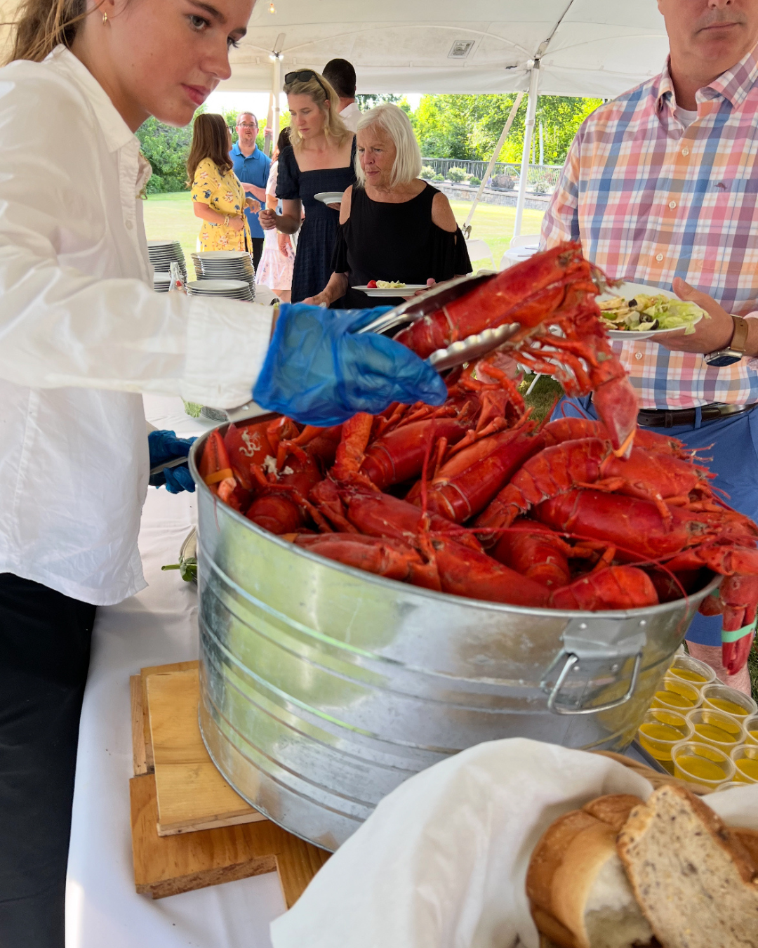 A woman in a white shirt and blue gloves serving cooked lobster from a large metallic tub at an outdoor buffet. People are in line with plates, and there is bread and small cups of sauce on the table. The setting is outdoors under a canopy, with gree
