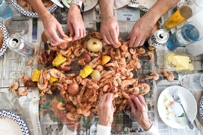 Overhead view of a seafood boil with shrimp, corn, potatoes, and lemons spread on newspaper; people are serving themselves with drinks nearby.