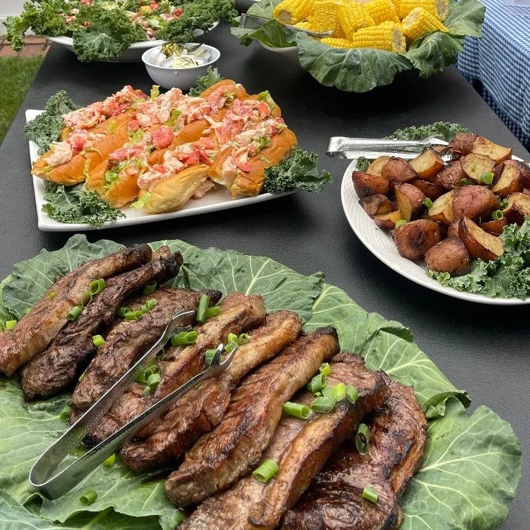 Assorted grilled meats and vegetables on a black table, including ribs garnished with chopped green onions, roasted potatoes, corn on the cob, and various salads.