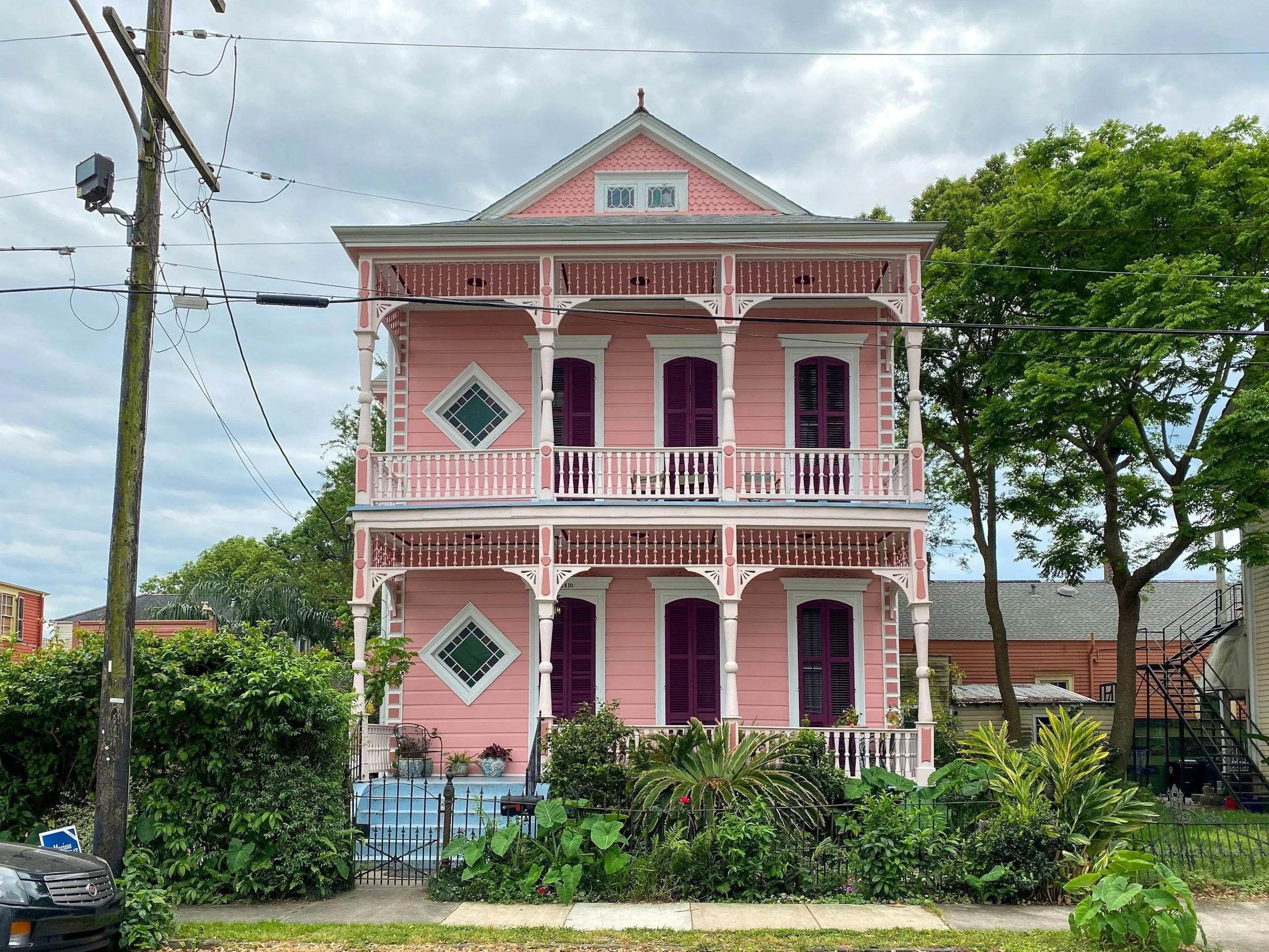 Pink three-story house with purple shutters and ornate white trim, front garden with plants, fence, and a small pool, overcast sky