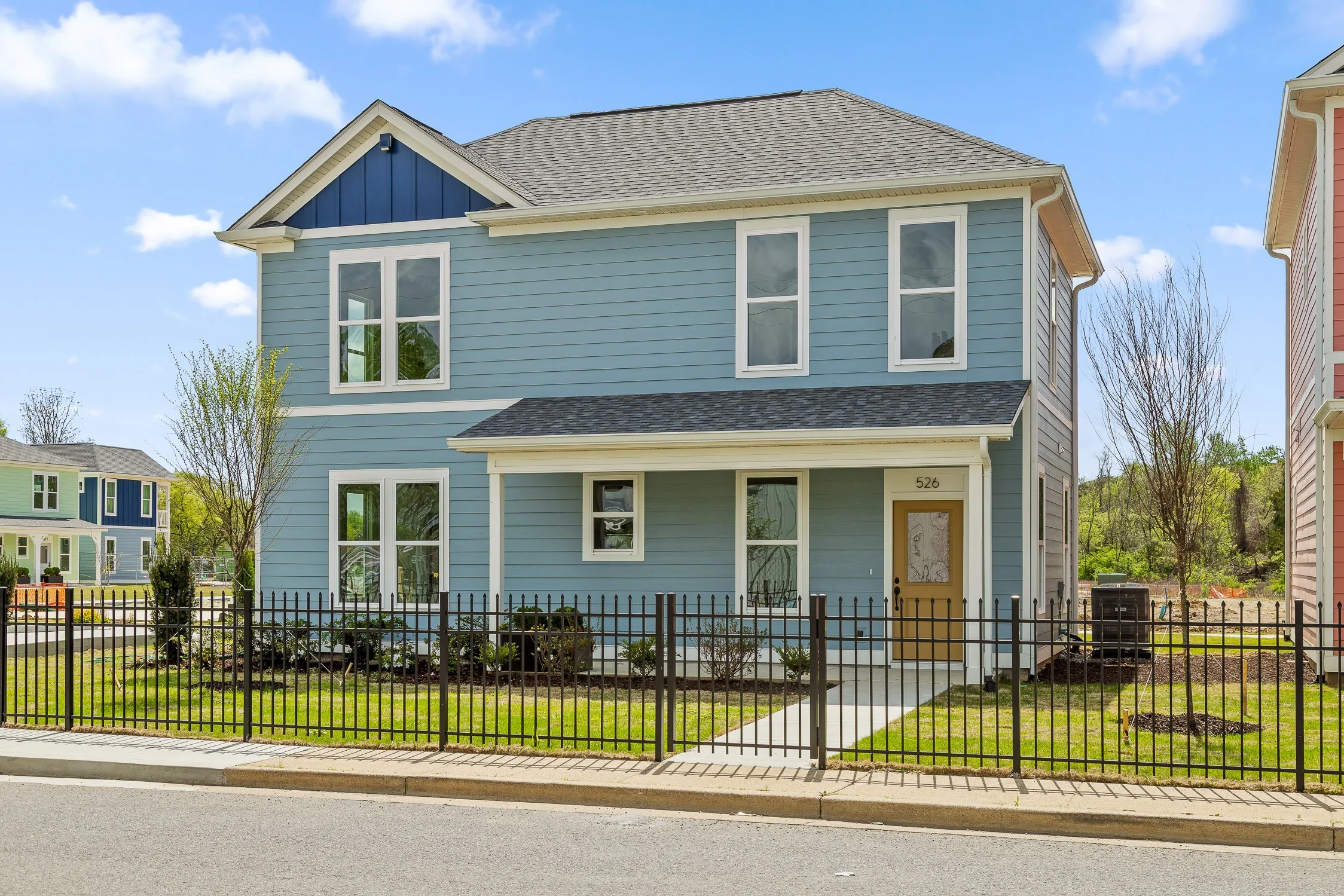 A two-story blue house with white trim and gray roof, fenced front yard, paved walkway leading to the front door, with neighboring houses visible in the background under a partly cloudy sky.