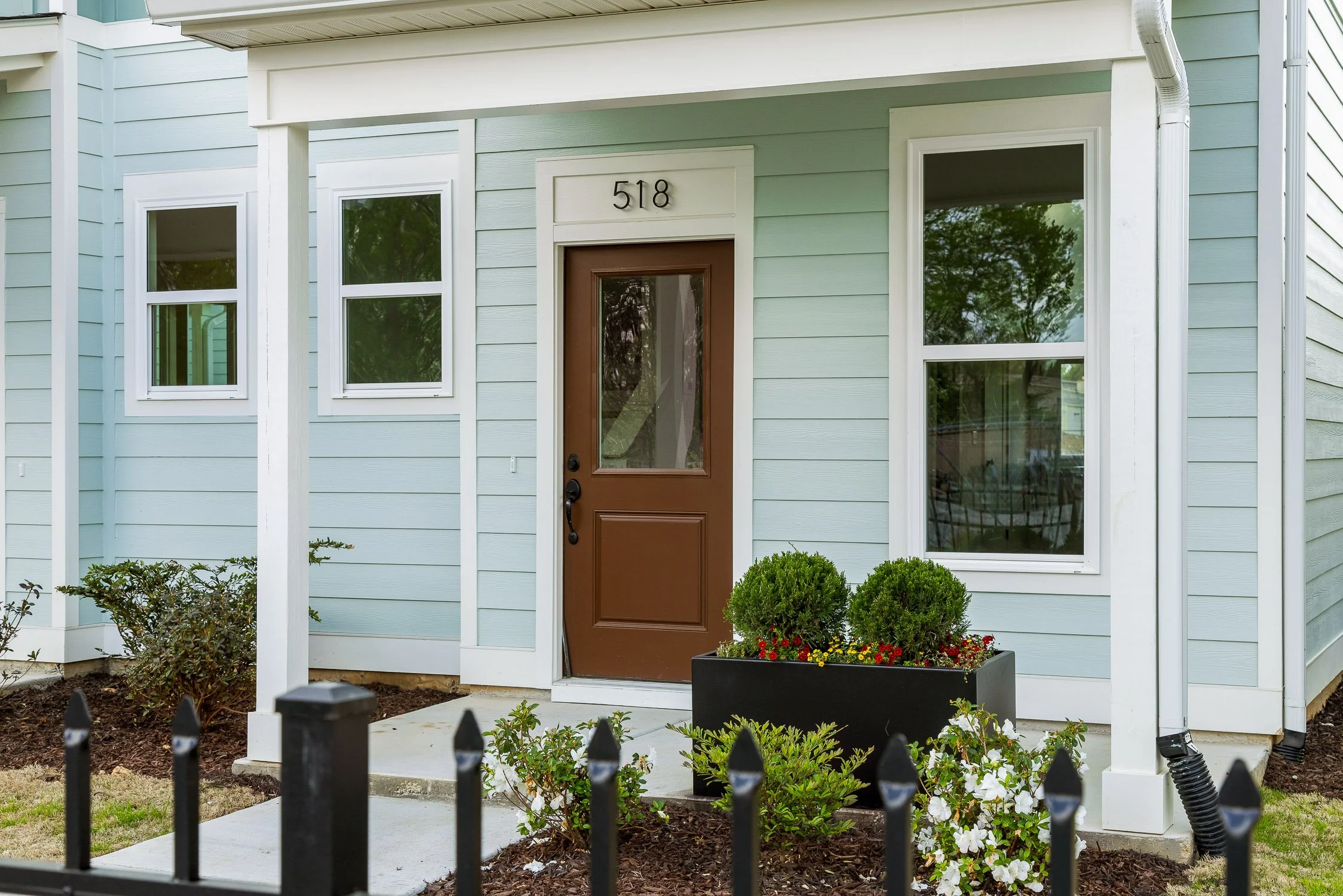Front view of a house with light blue siding, a brown door, and white framed windows. The house number 518 is above the door. There is a small garden with bushes and a large black planter with flowers in front of the house. A black metal fence is visible in the foreground.