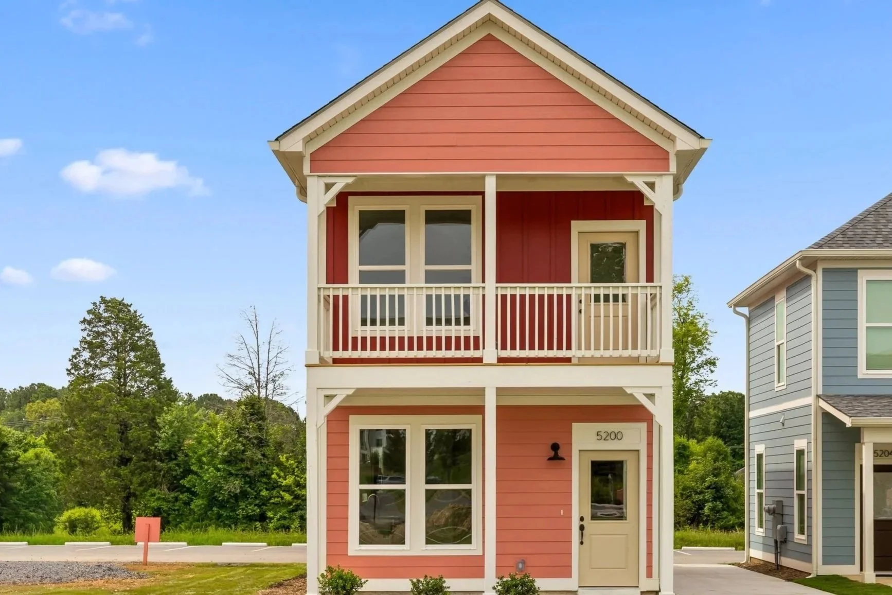 Three-story pink house with a front balcony, white trim, and a beige door number 5200, located in a suburban neighborhood with a neighboring blue house to the right and greenery in the background.