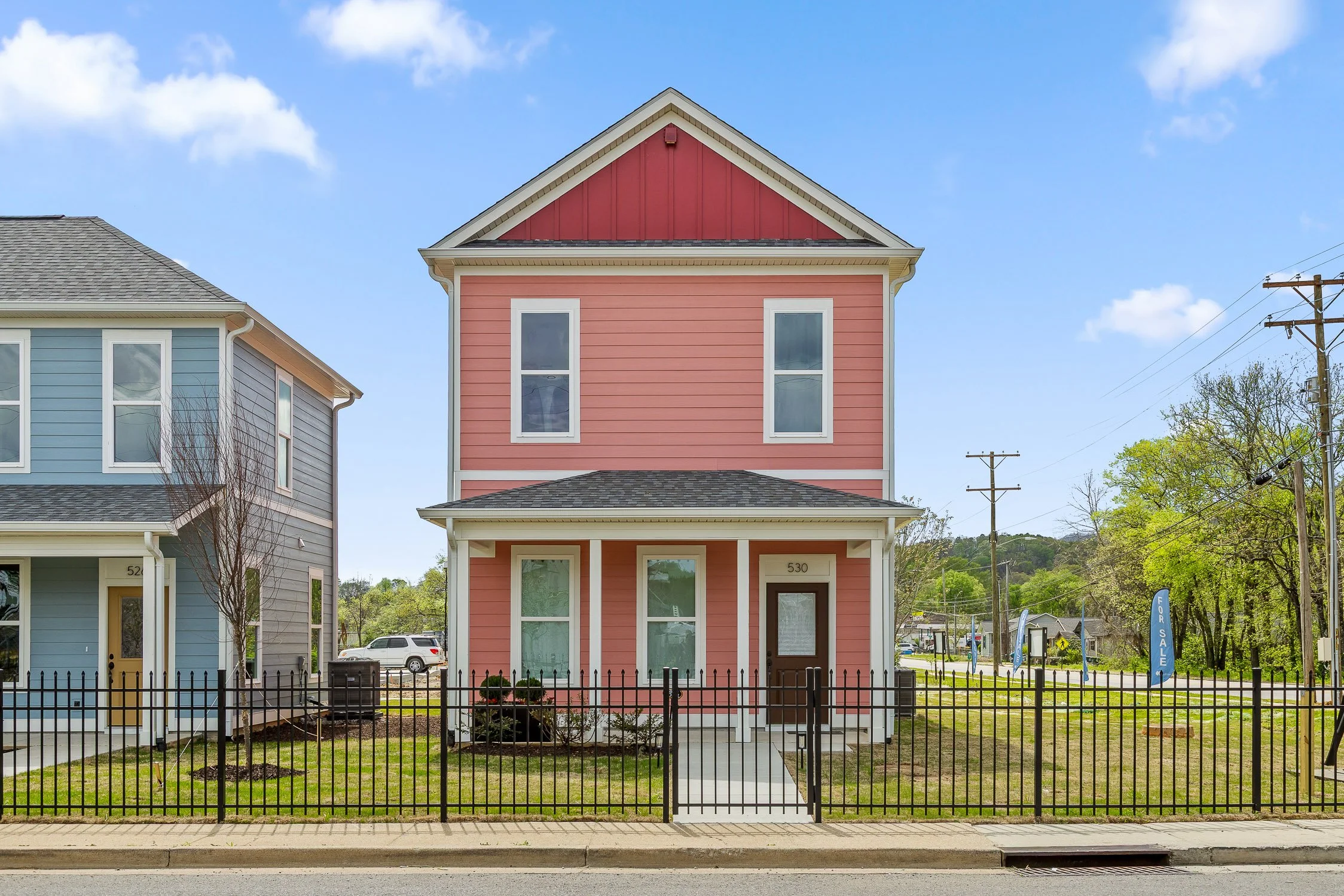 Pink three-story house with a small front yard, black metal fence, and a sidewalk, adjacent to a gray house on the left, under a blue sky with scattered clouds.