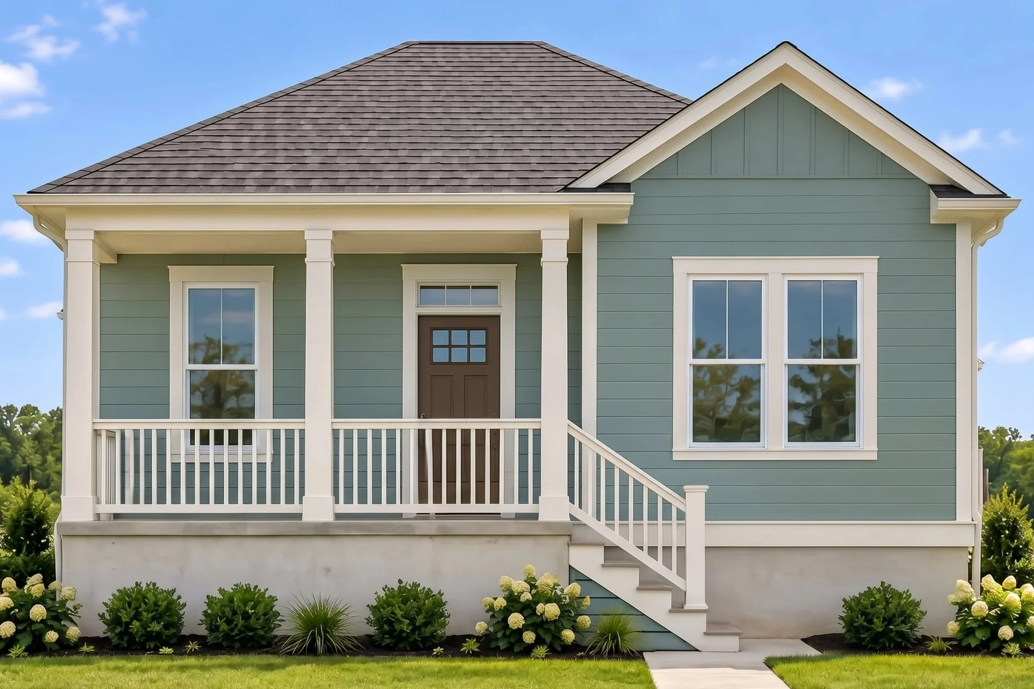 Front view of a light blue house with white trim and a staircase leading to a porch, outdoor plants and grass in the yard, and a clear blue sky.
