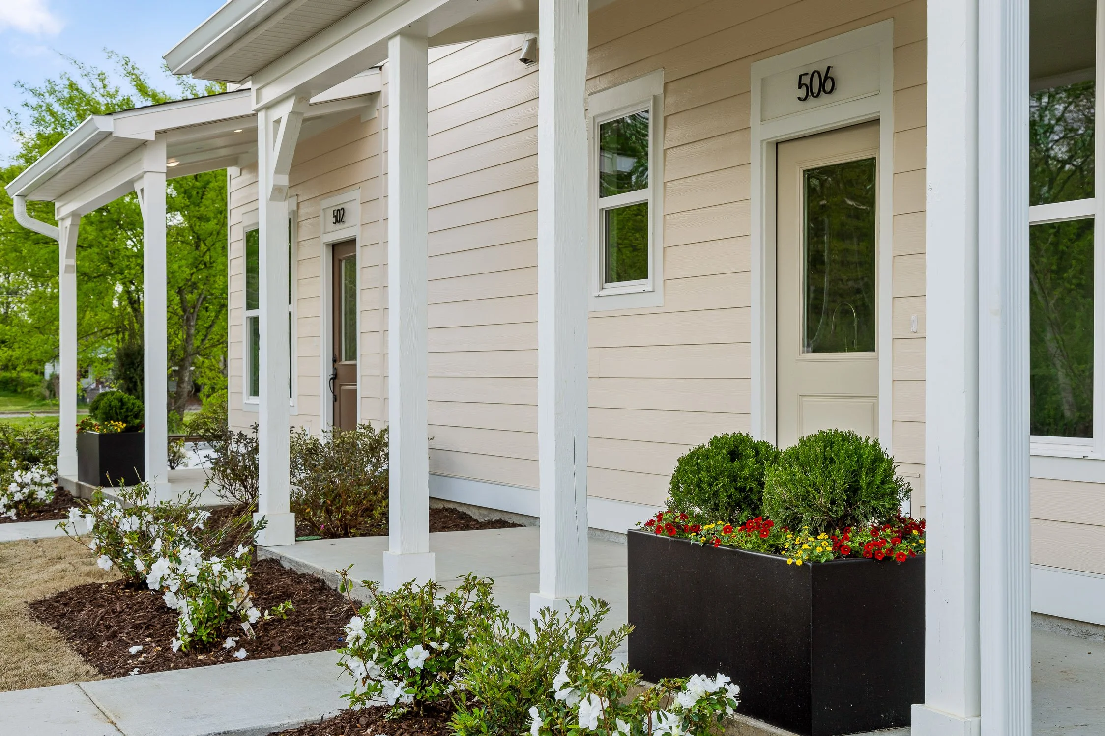 Front of a beige apartment building with two doors, numbered 502 and 506, each with a small window and surrounded by small landscaped gardens with bushes and flowers.