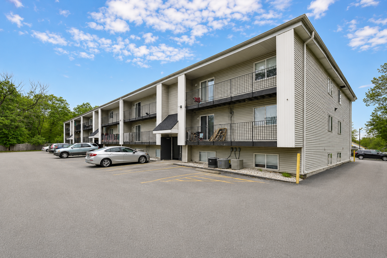 Three-story apartment building with balconies and parked cars in front, under a partly cloudy sky.
