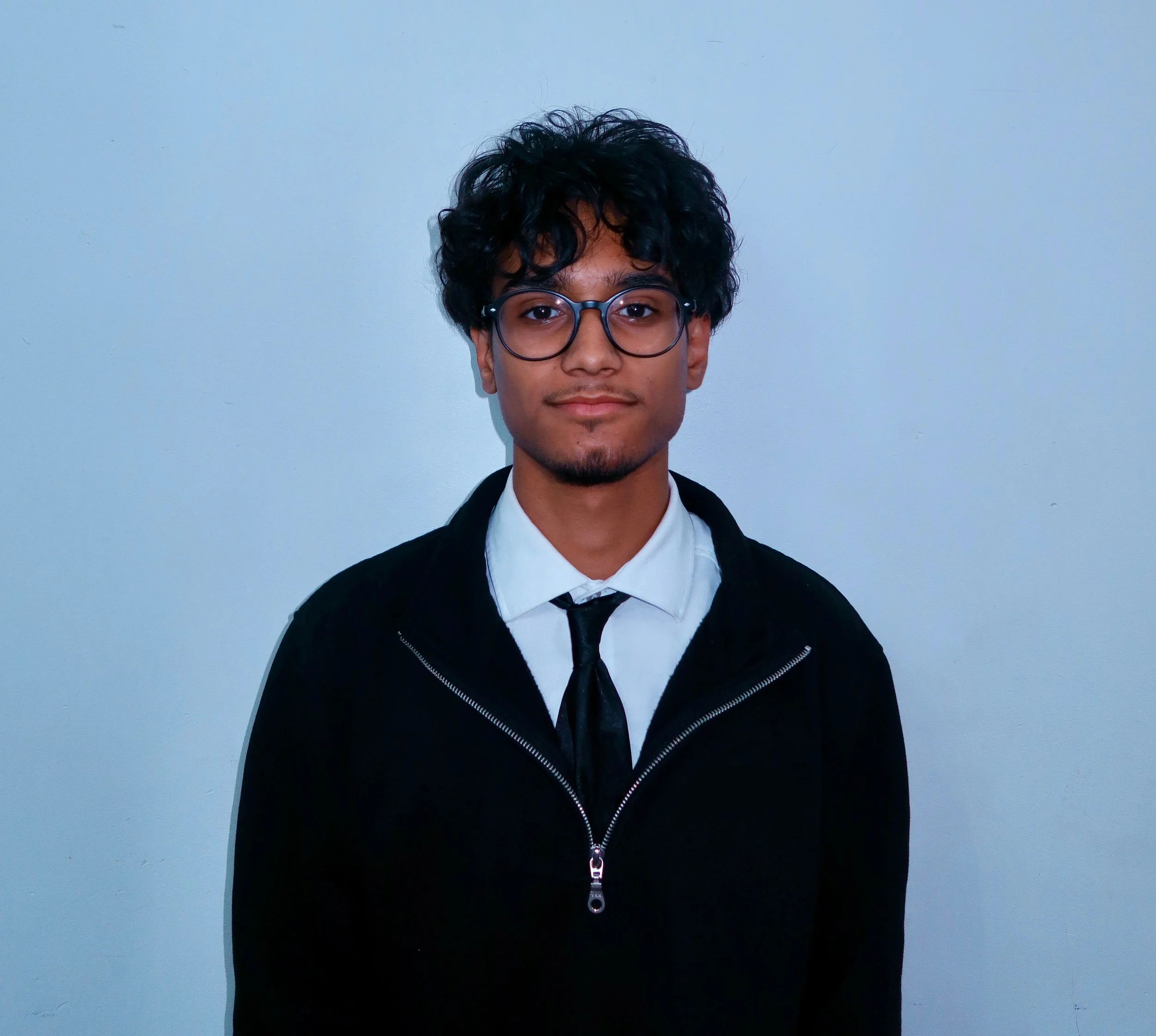 Young man with black curly hair, glasses, wearing a white shirt, black tie, and black jacket, standing against a plain light blue wall.