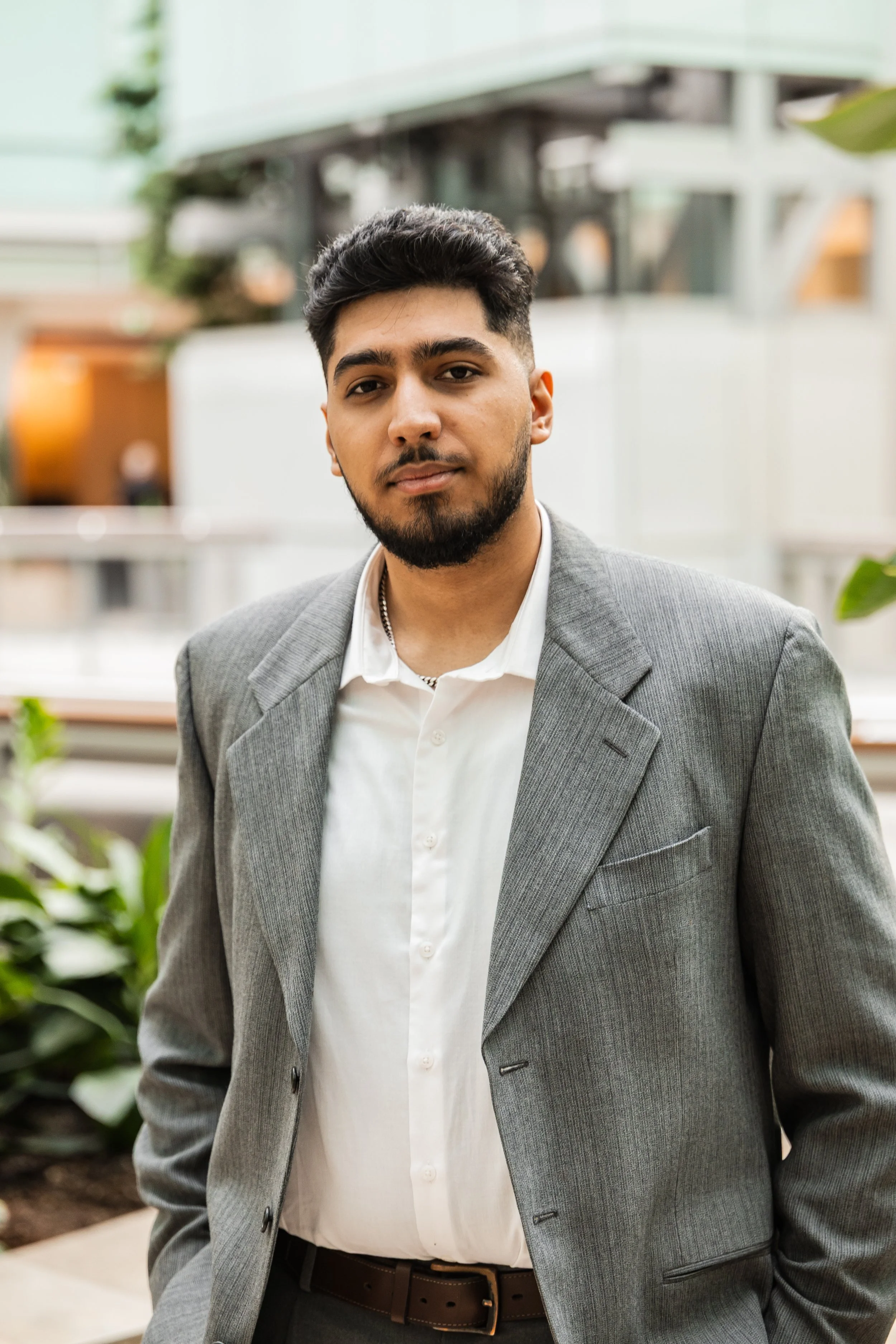 A young man with a beard and short dark hair wearing a gray suit jacket and white shirt standing outdoors with a blurred modern building in background.