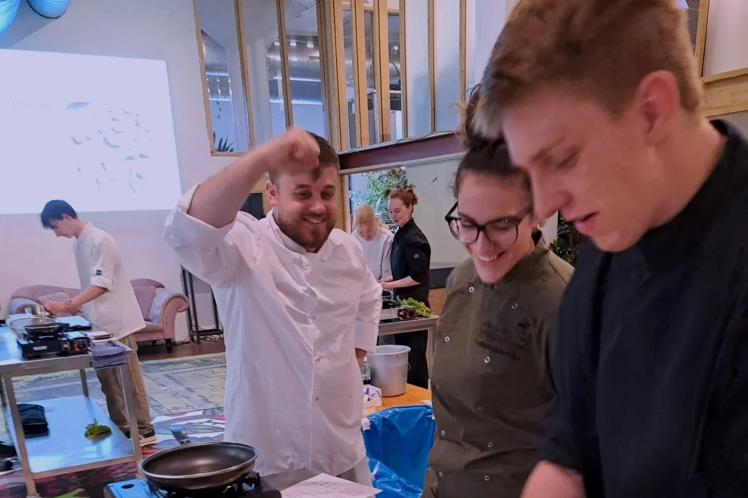 Interieur van een keuken of cafetaria met werkbladen, stoelen, keukengerei en apparatuur, met grote ramen dat een industriële ruimte toont.