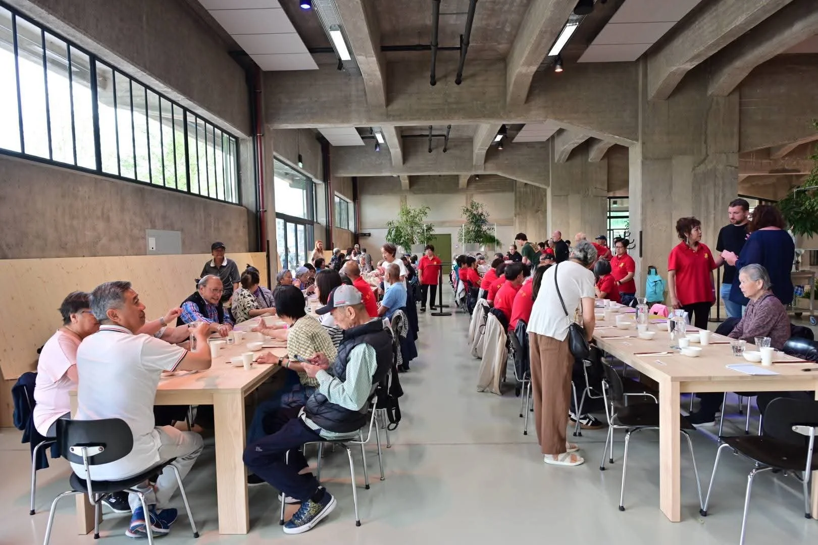 Een grote groep Chinese vrijwilligers mensen zit aan lange tafels in een industriële, moderne ruimte met hoge plafonds en grote ramen. Sommige mensen wachten, andere praten of eten. Vrouwen in rode uniformen staan en praten met gasten. leeftijd 65+