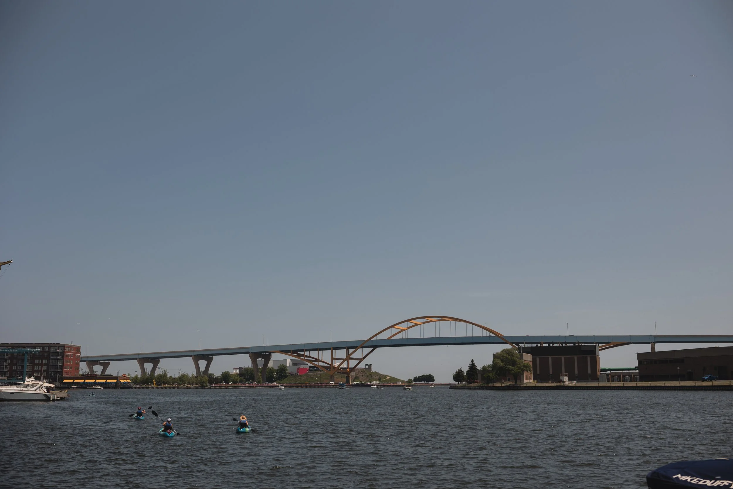 View of a river with three kayakers paddling, a boat on the water, and a bridge with a curved arch in the background, under a clear blue sky.