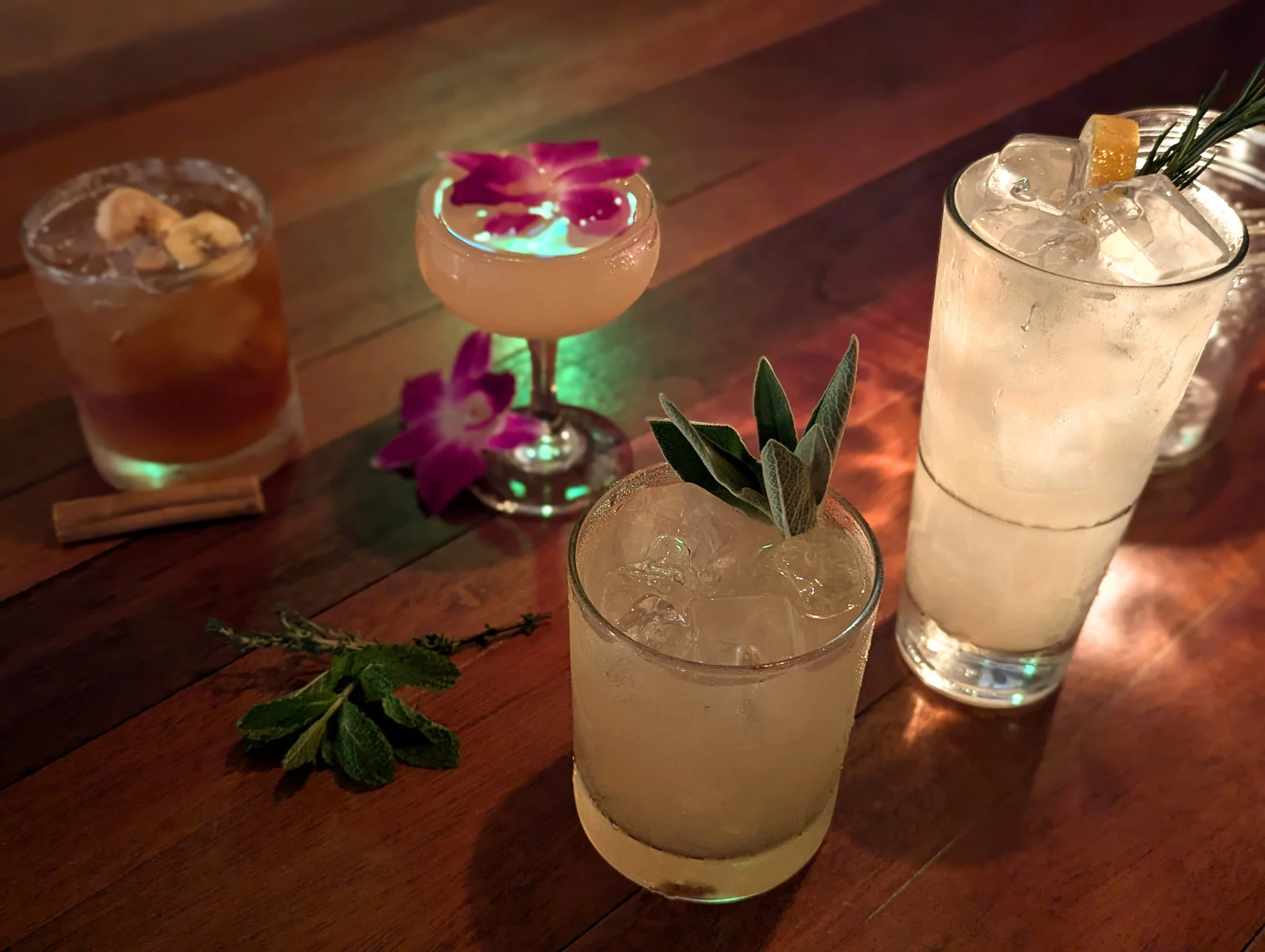 Three cocktails on a wooden table with fresh herbs and flowers for garnish, illuminated by warm lighting.