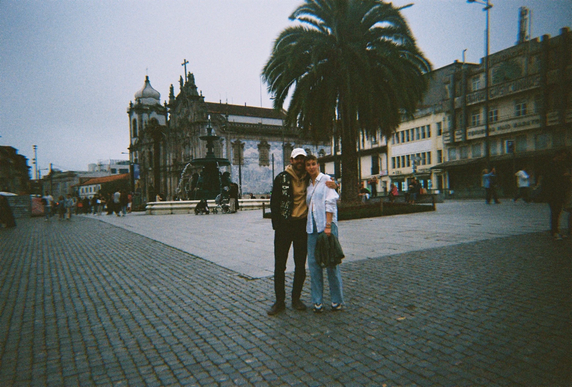 A couple standing together on a cobblestone plaza in Porto, Portugal, with a historic church and palm tree in the background. After planning their new website of theit family company LUCCHINI MOON that offers leisure clothing and dainty jewelry.