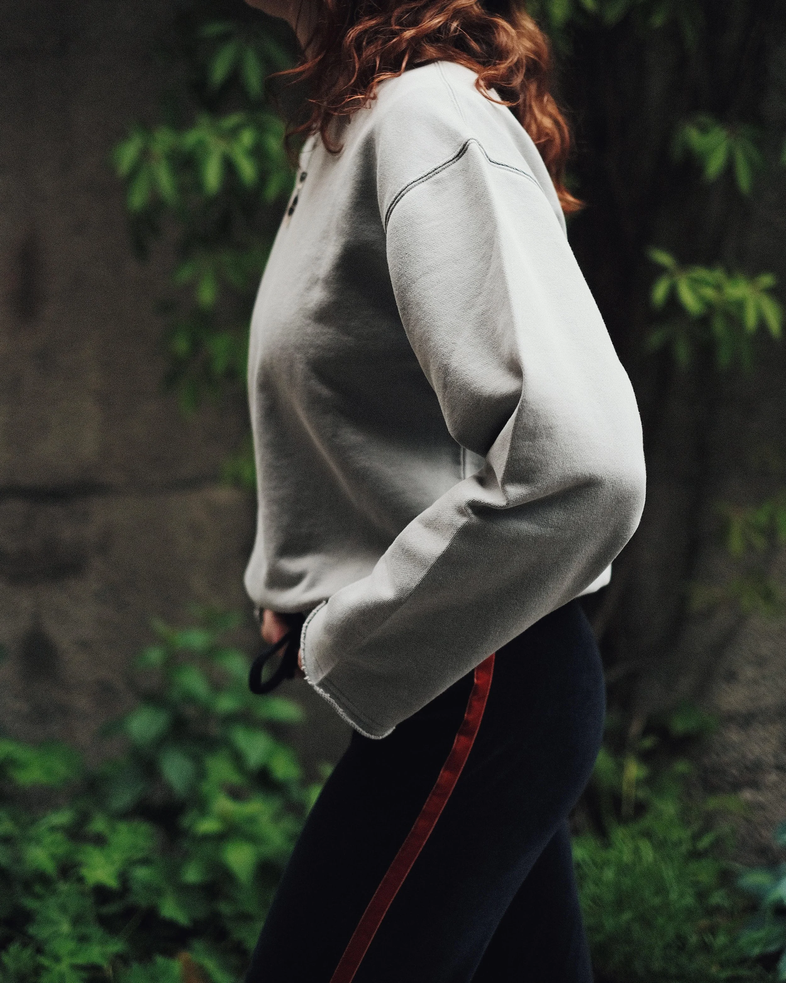 A person with curly red hair wearing a light gray sweatshirt and dark pants with a red stripe, standing outdoors near green foliage.