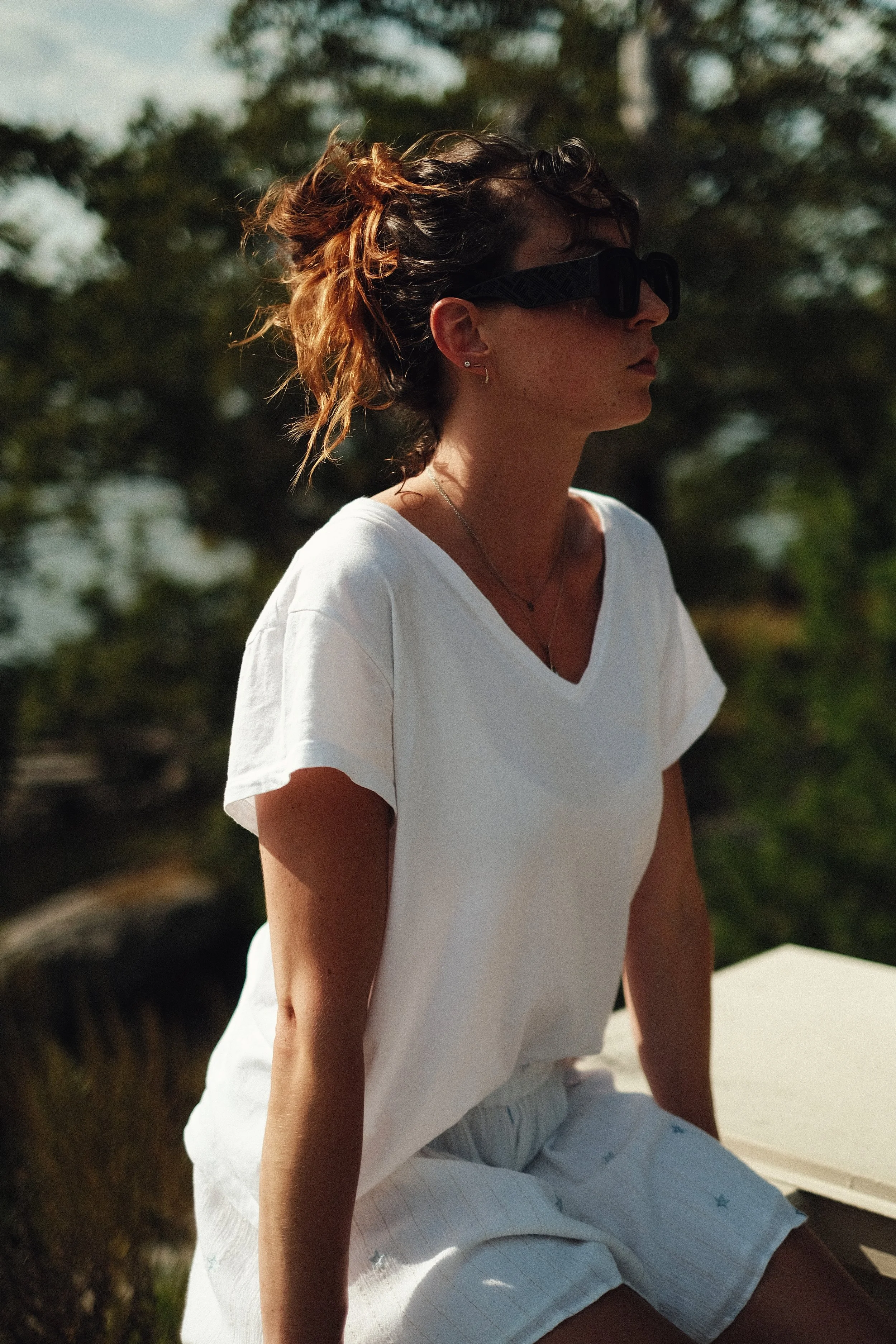 Woman wearing sunglasses, white t-shirt, and shorts sitting outdoors on a sunny day with trees in the background.
