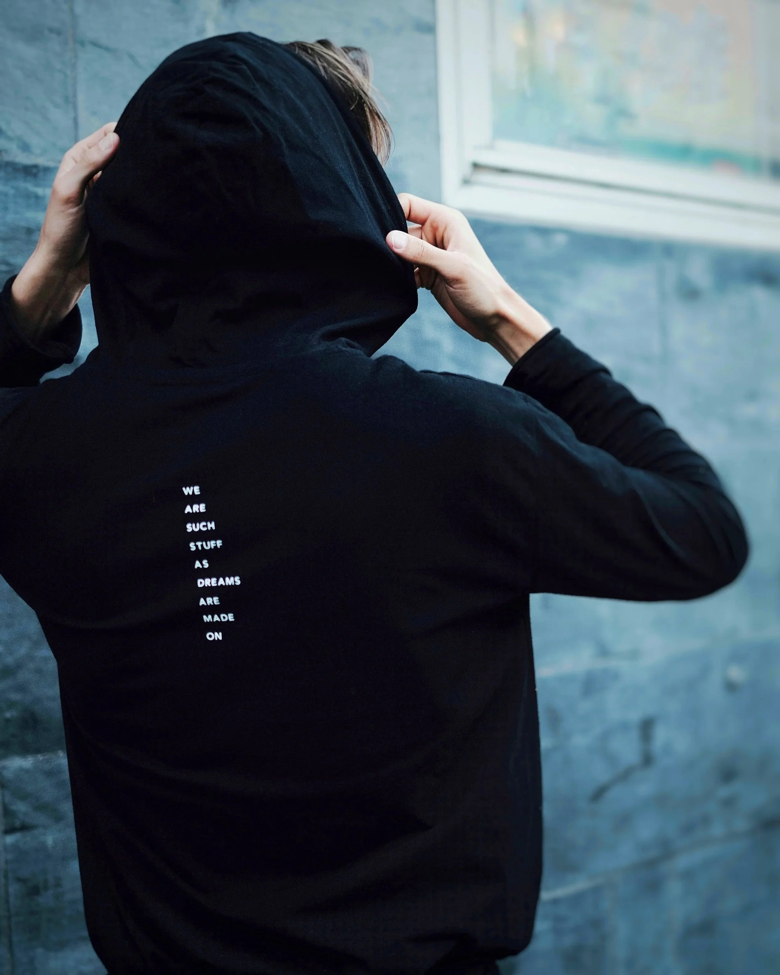 Person wearing a black hoodie in 100% organic cotton with white Shakesperian text on the back, holding the hood over their head, standing in front of a window in Stockholm with a concrete wall background.