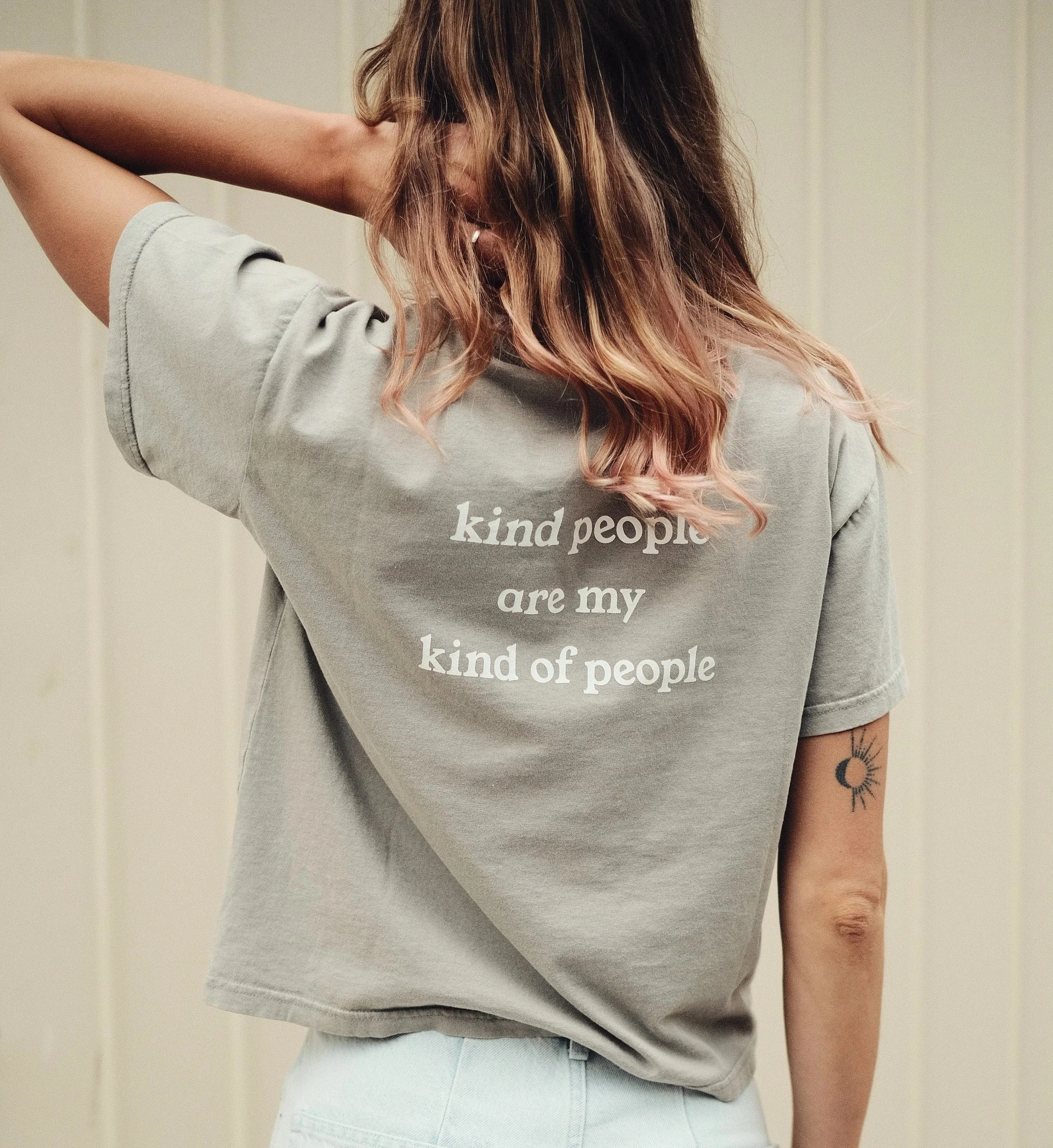 A person with wavy hair wearing a gray t-shirt that says "kind people are my kind of people" on the back. The person has a tattoo of a sun and moon on the left arm.