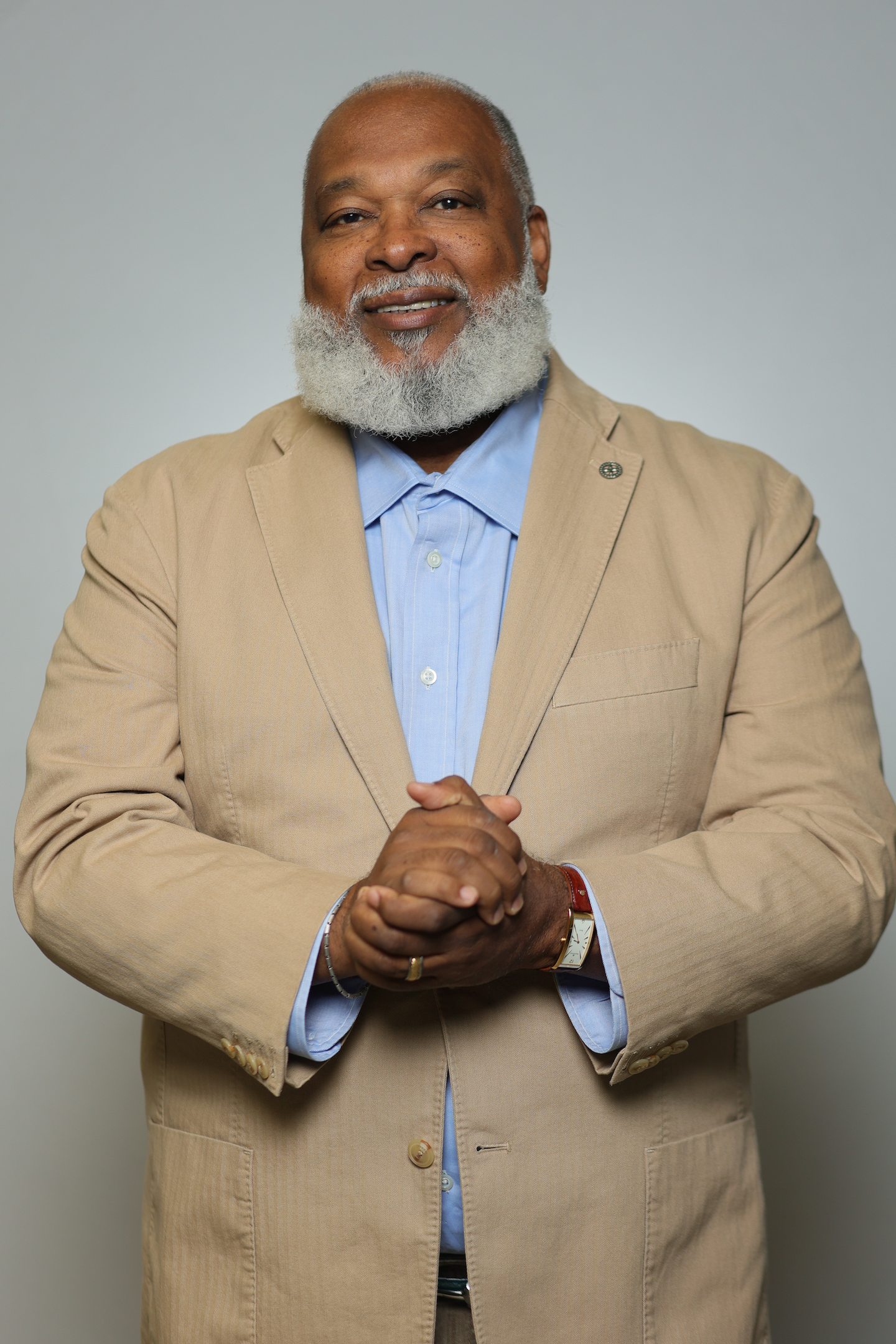 A middle-aged man with dark skin, a gray beard, and short gray hair, wearing a beige suit jacket over a light blue dress shirt, standing with hands clasped in front of him against a plain gray background.