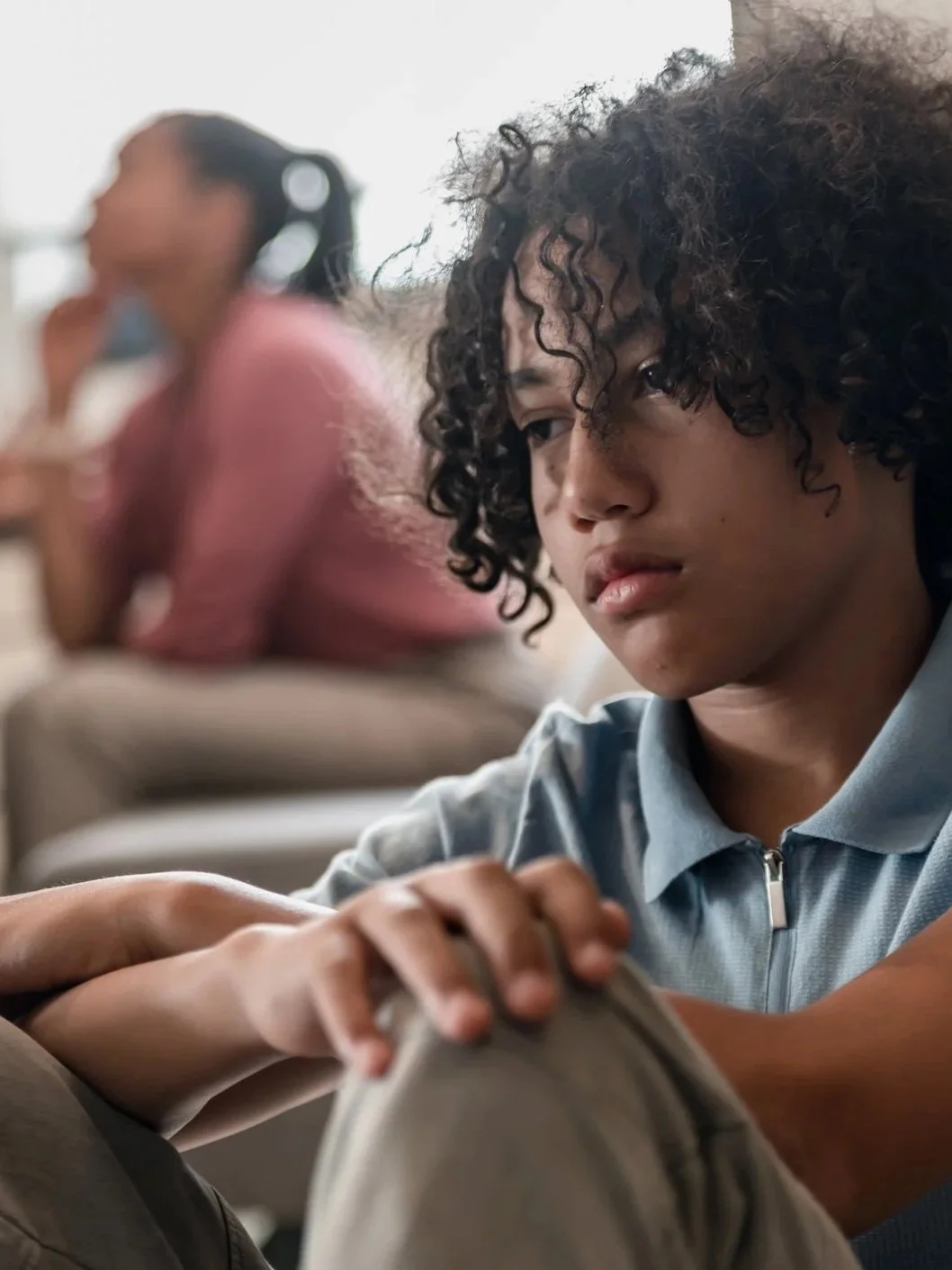 A young person with curly hair sitting with arms resting on their knees, looking down thoughtfully. In the background, a woman in a pink sweater with glasses is blurred, sitting and resting her chin on her hand.