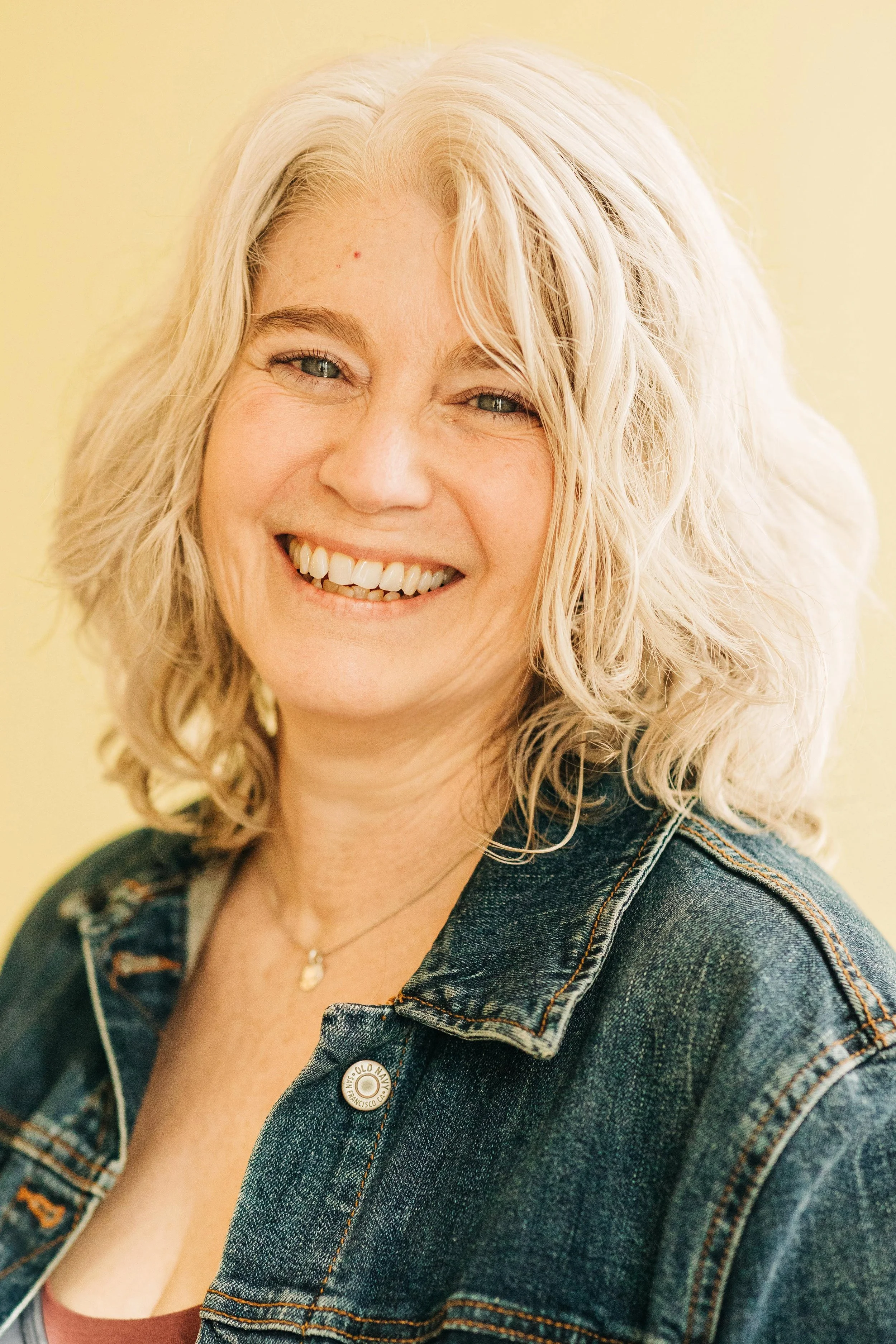 Smiling woman with blonde hair wearing a jean jacket against a yellow background.