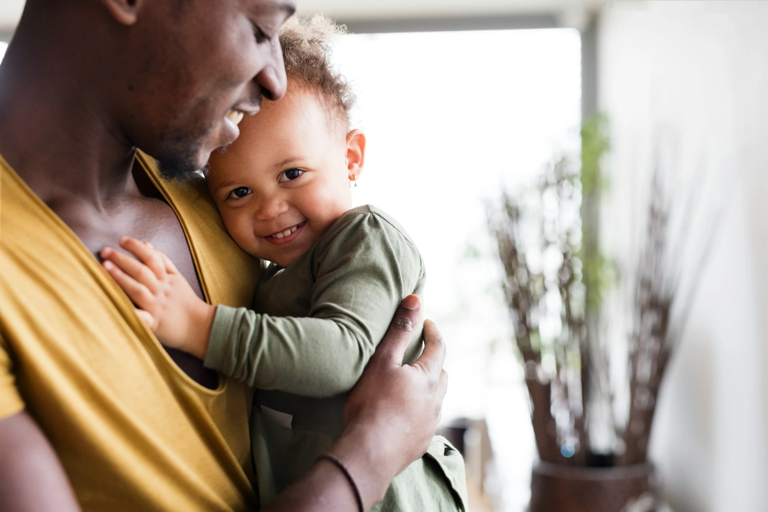 A man holding a smiling young girl close, both inside a room with bright natural light and a blurred plant in the background.