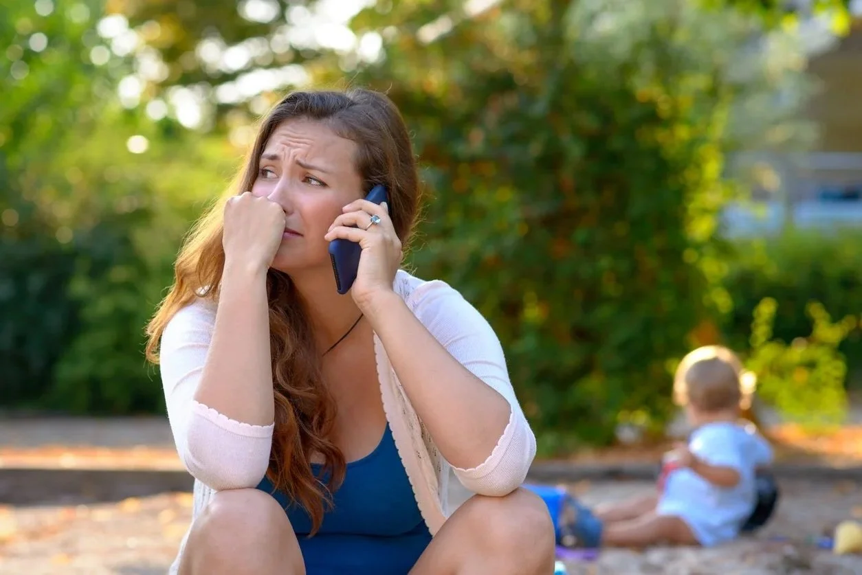 A woman with long brown hair sitting outdoors, holding a phone to her ear and appearing worried or anxious, with a young child sitting on the ground in the background.