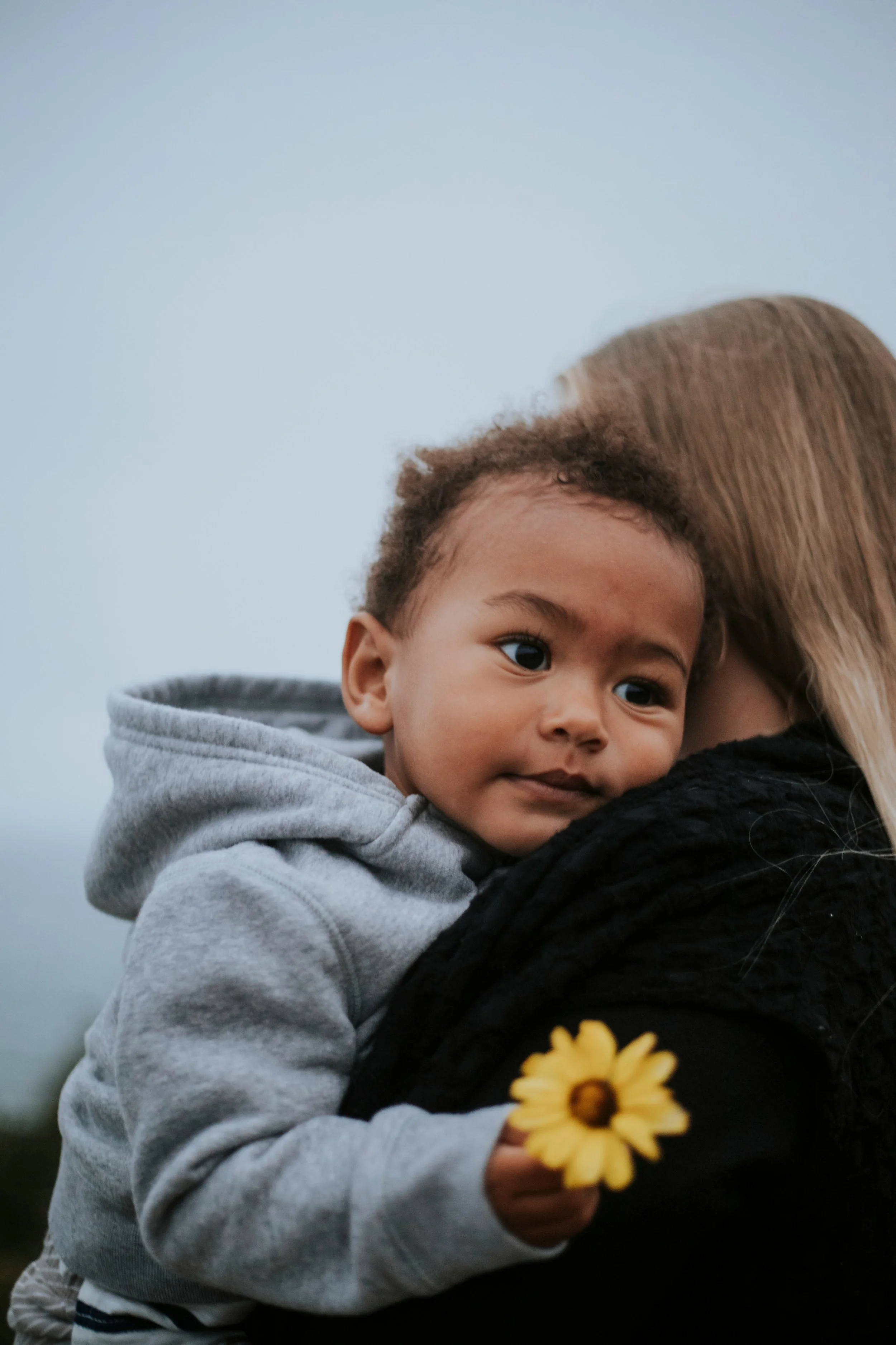 A young child with curly hair, wearing a gray hoodie, is resting their head on an adult's shoulder, holding a yellow flower in hand.