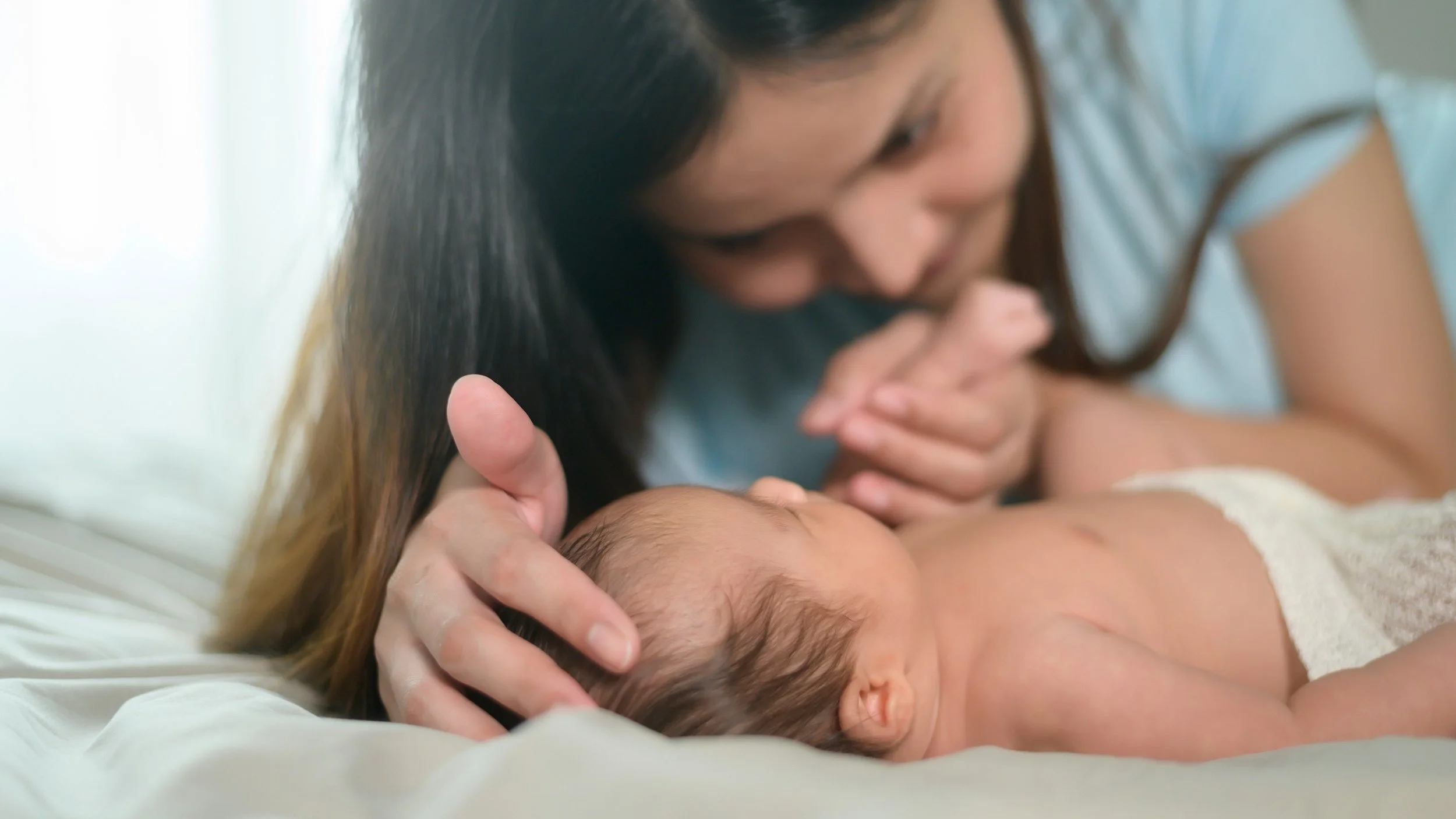 A woman gently kisses a sleeping newborn baby on the forehead while lying on a bed.