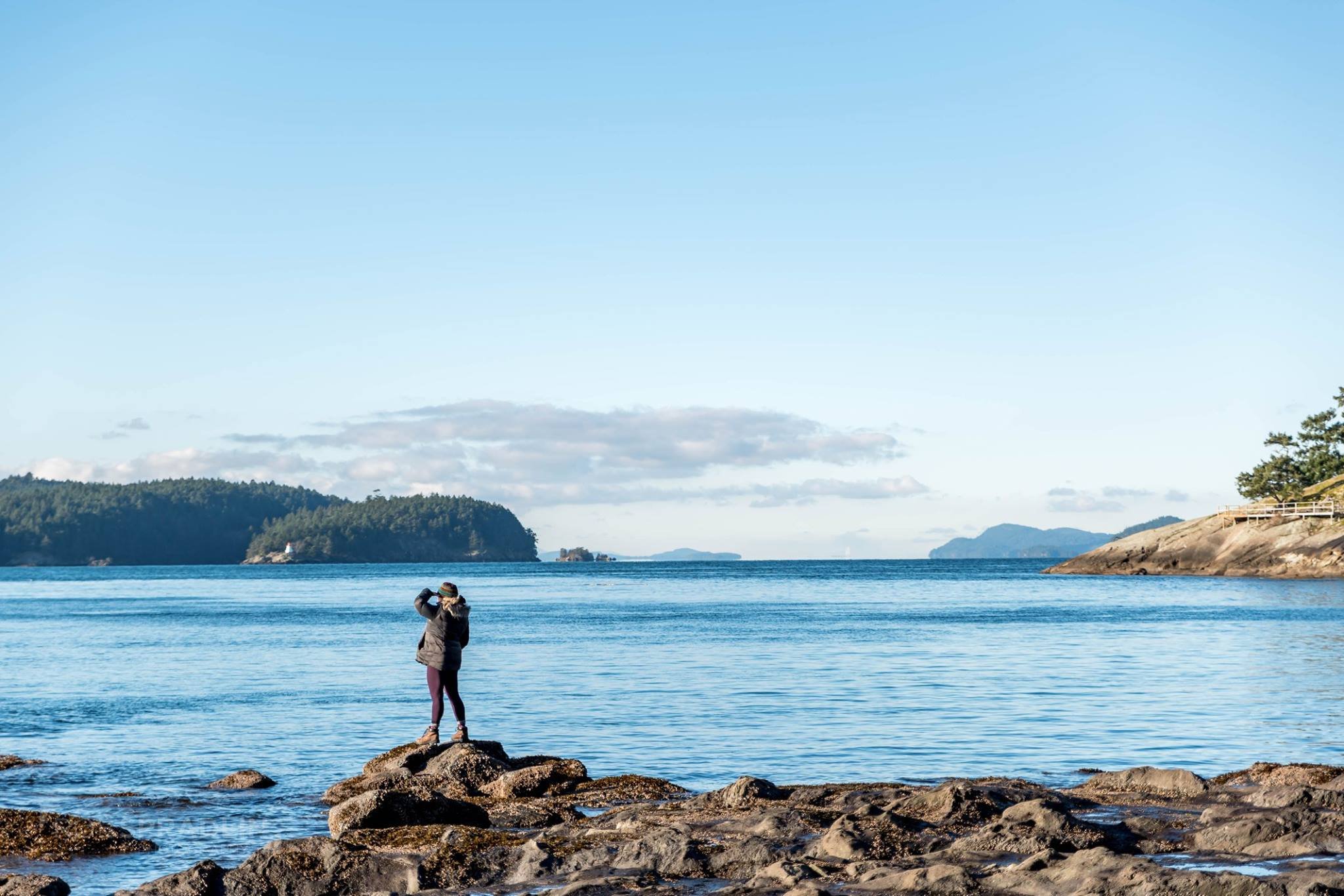 Person standing on rocks at the edge of the water, taking a photo of the ocean and distant islands under a blue sky with some clouds.