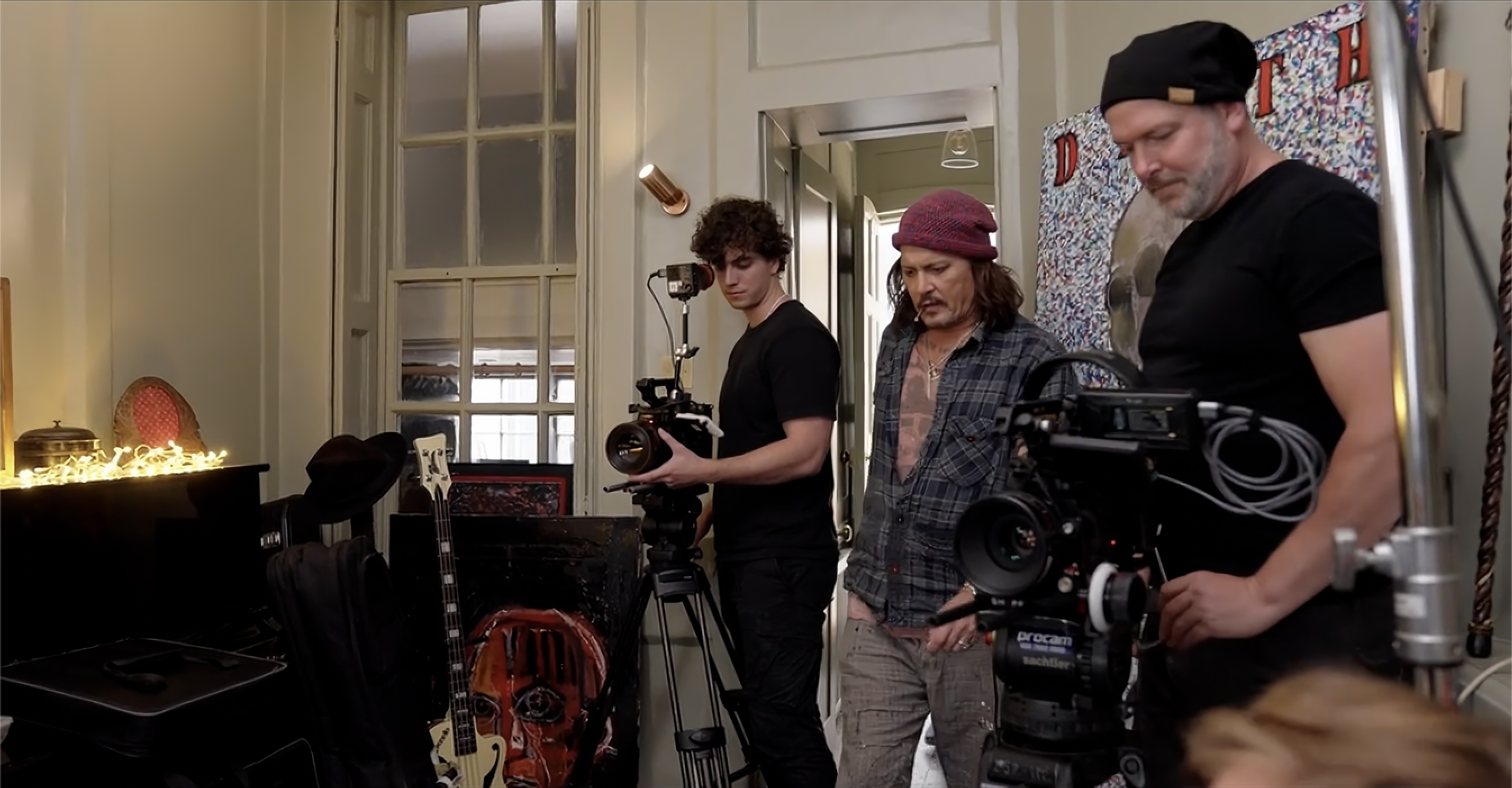 Three film crew members standing in a room with musical instruments and a window, preparing for filming.