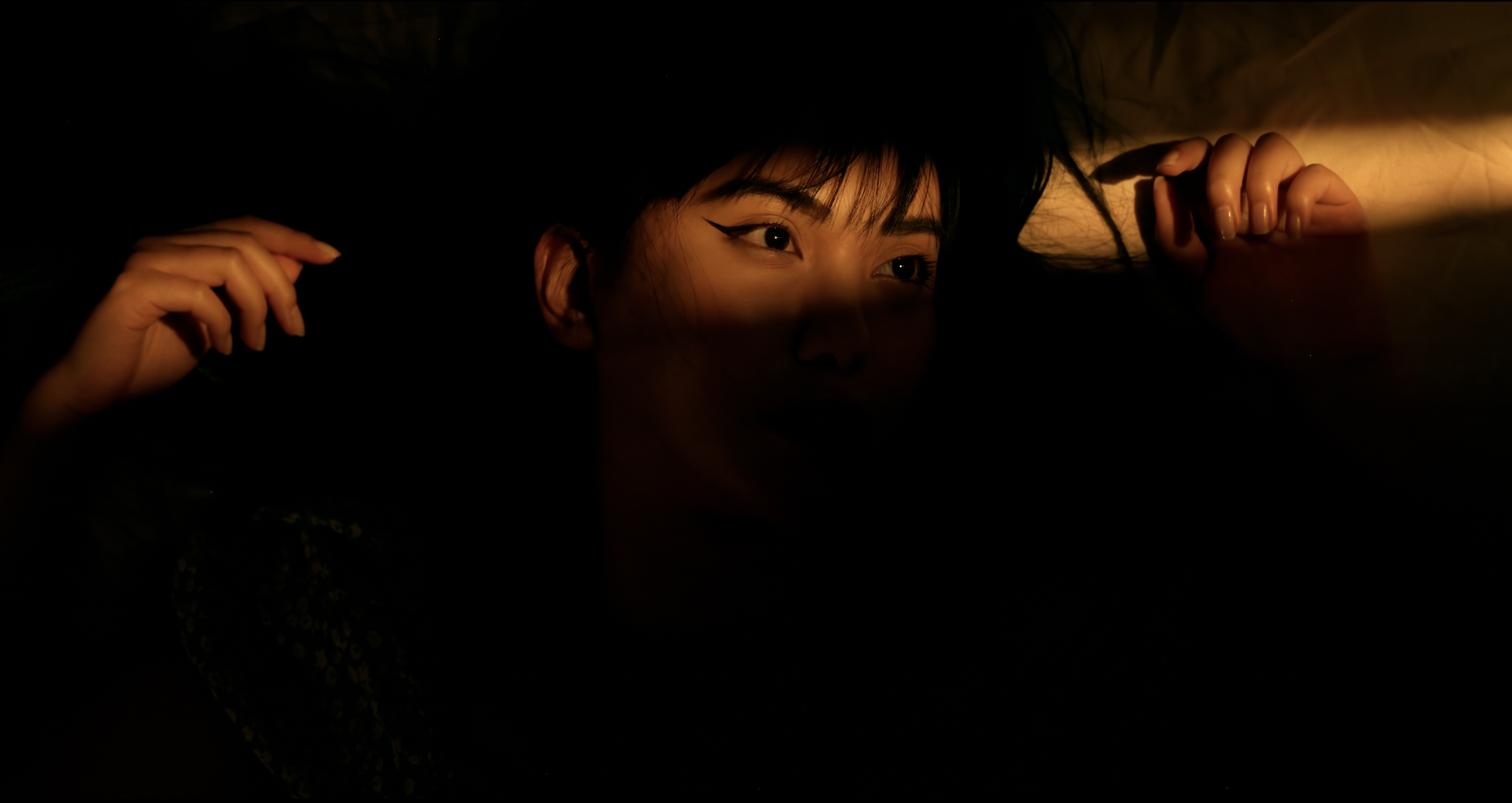 A close-up of a person's face partially illuminated by warm, low light, with hands gently holding long, dark hair, and only part of their face visible in shadow.