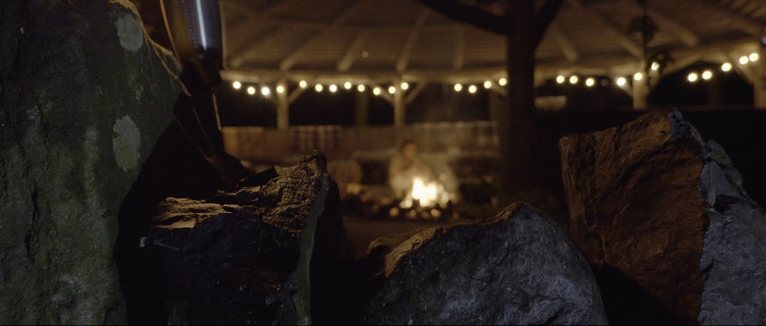 Rocks in the foreground with a fire pit and a person sitting near it in the background under a thatched roof with string lights.