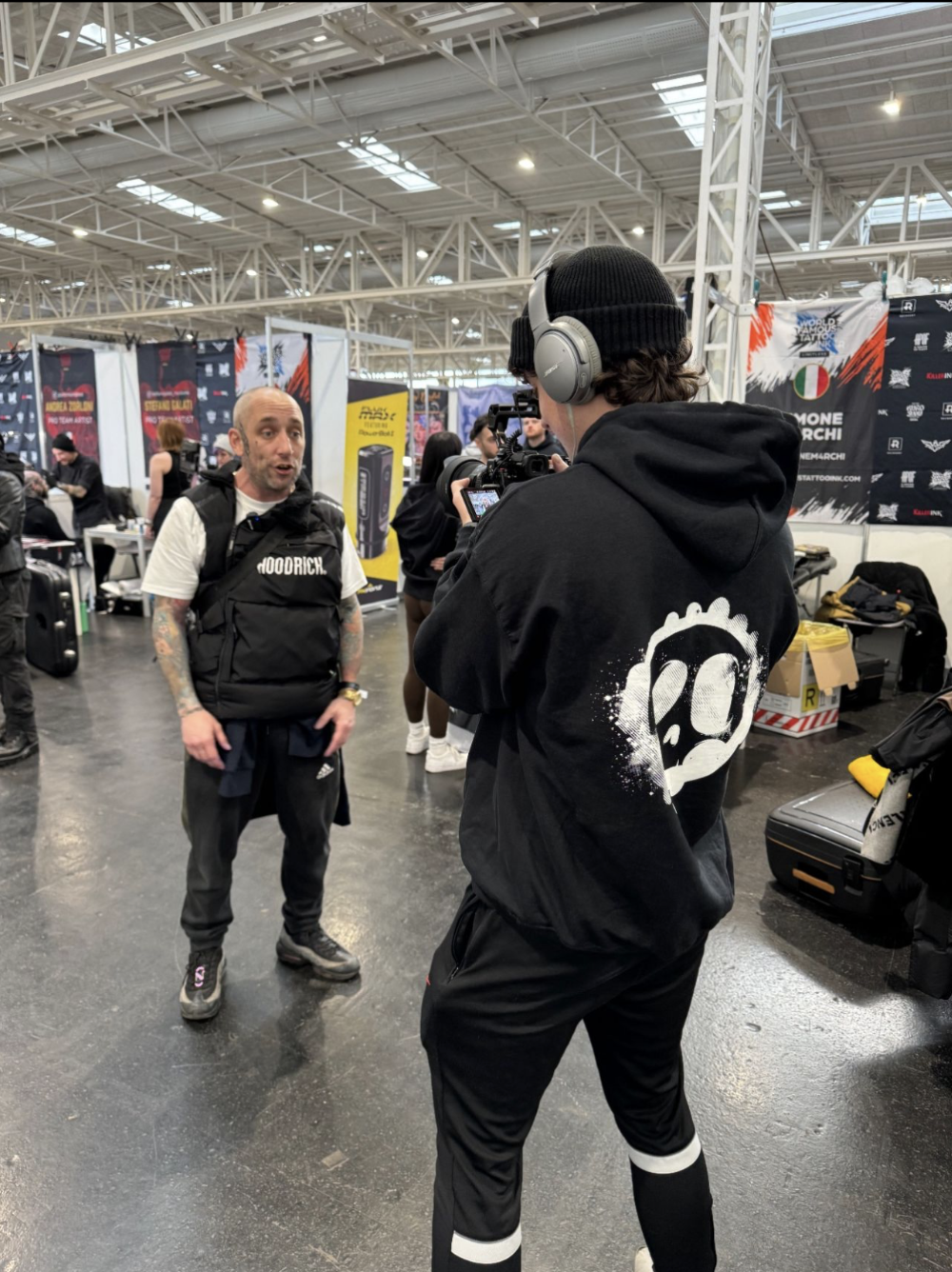 Two men are having a conversation at an indoor event. One man appears to be speaking and is wearing a black vest and tattooed arms. The other man, seen from behind, is wearing headphones, a black hoodie with a skull graphic, and is holding a handheld device. There are multiple booths and banners in the background.