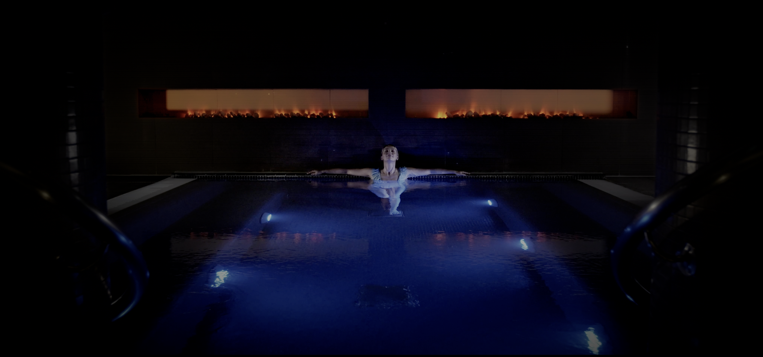 A woman floating in a dark indoor pool with her arms outstretched, illuminated by blue lights, and a fiery fireplace in the background.