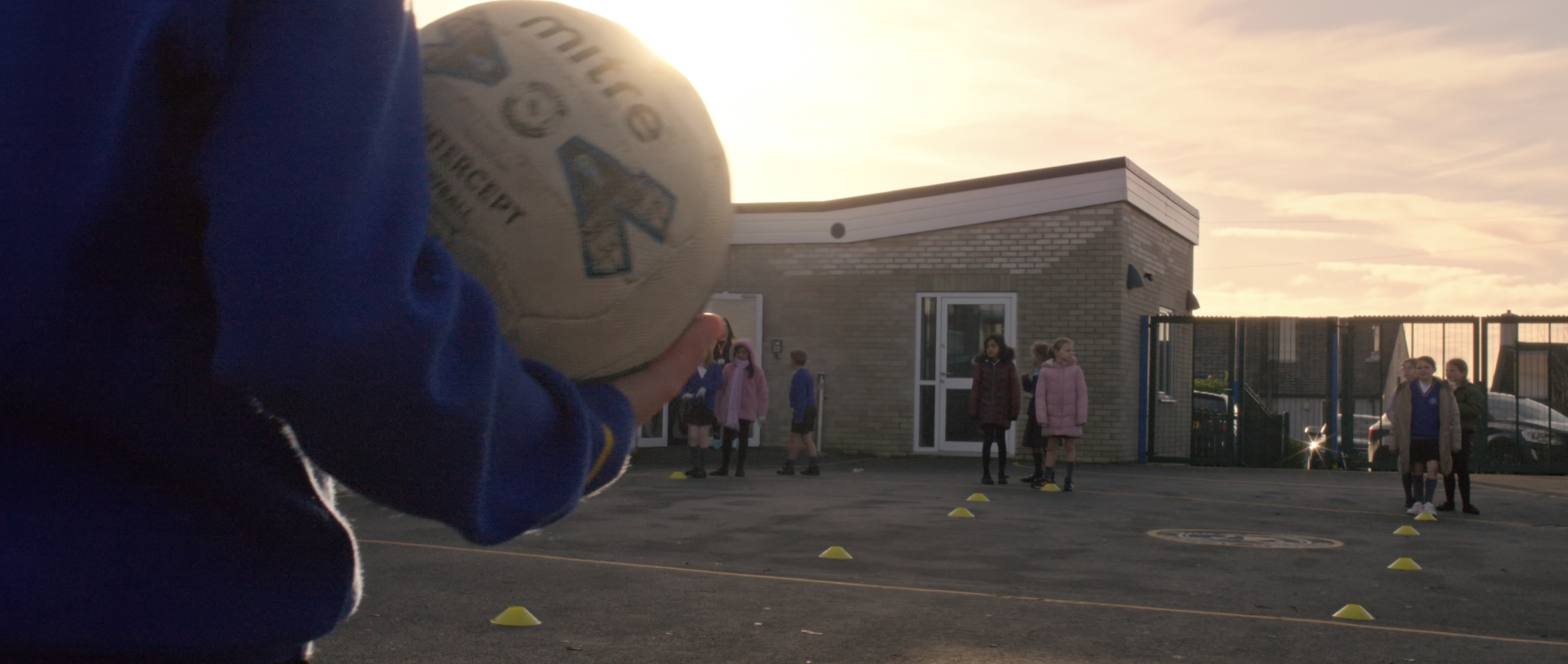 A person holding a netball with a schoolyard and children in the background during sunset.
