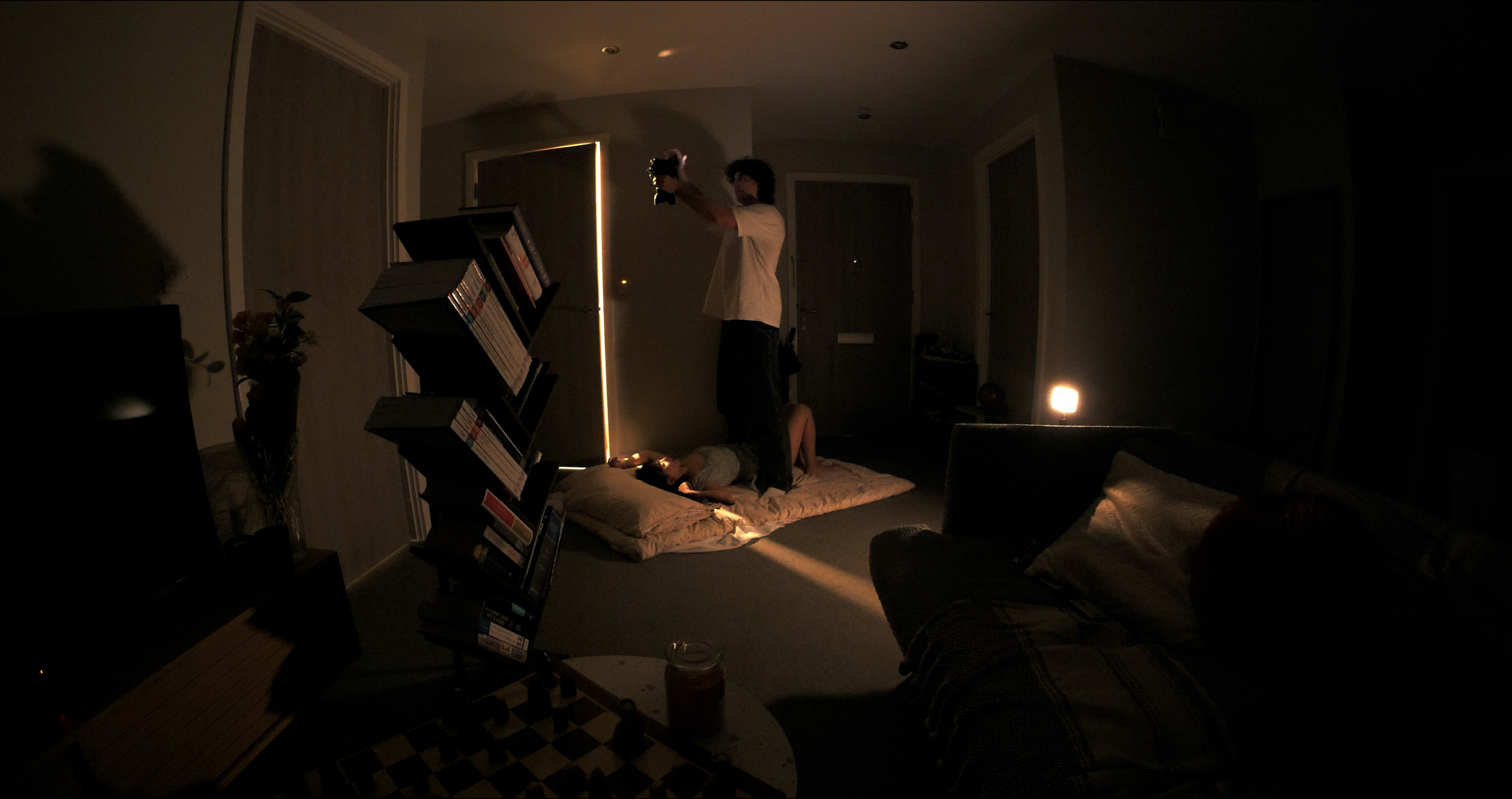 A dimly lit living room with two people on a mattress on the floor; one person is lying down while another stands, holding a camera. A bookshelf with books, a small table with a jar, and a couch with pillows are visible, with soft ambient lighting.