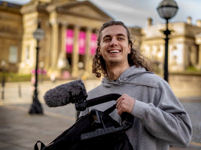 A young man with long hair smiling while holding a microphone, standing outdoors in front of a historic building.