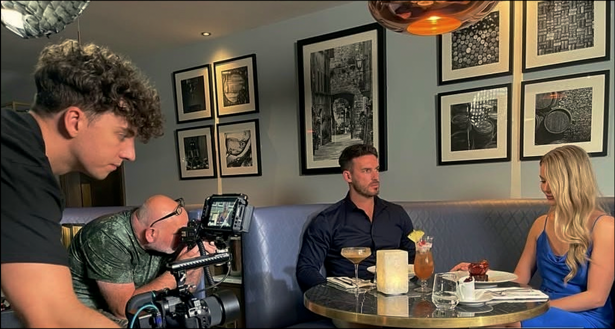 A film crew is shooting a scene at a restaurant with two actors, a man and a woman, sitting at a table with drinks and food. The man wears a dark shirt, the woman a blue dress. A cameraman and a director operate the equipment. The background features black and white framed artwork on the wall.