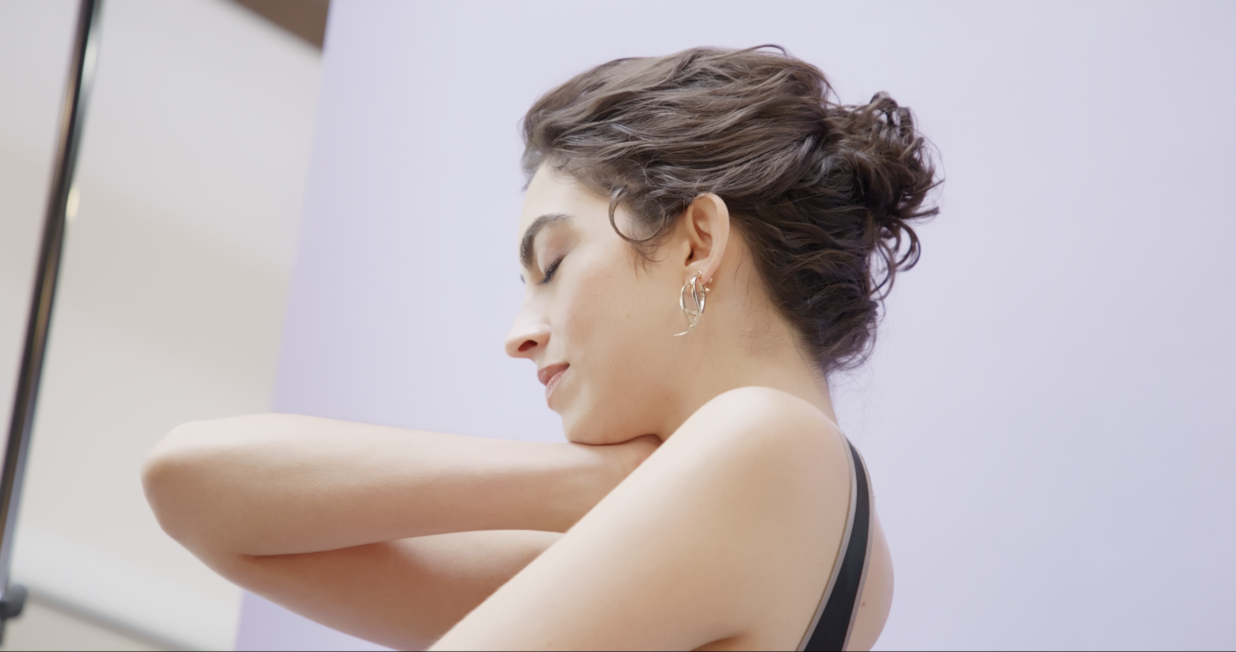 A woman with short, curly brown hair and earrings, resting her head on her forearm with closed eyes, against a plain light purple background.