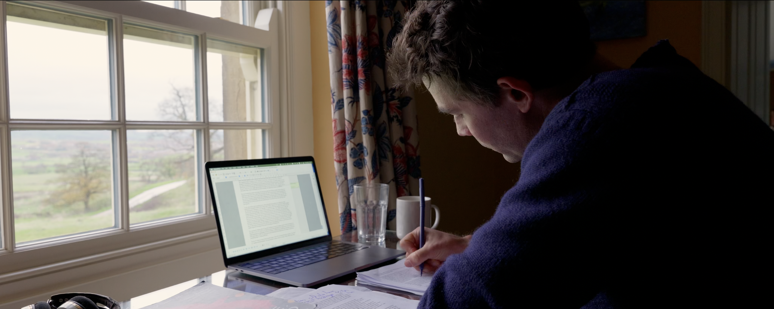 A person sitting at a desk near a window, working on paperwork with a laptop, a glass of water, and a mug on the desk.