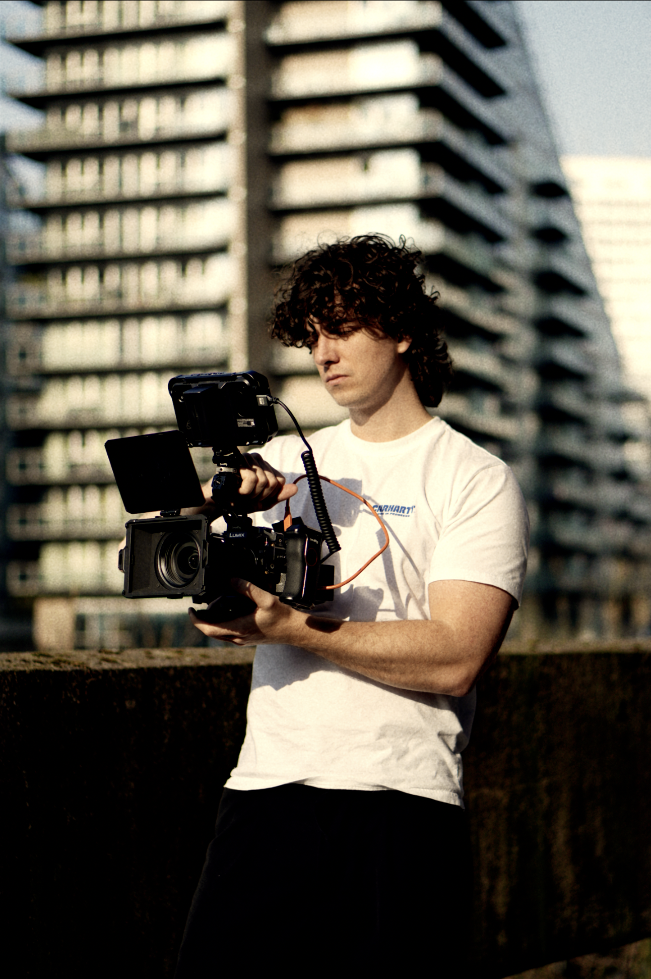 A man with curly hair wearing a white t-shirt is holding a professional video camera outdoors in front of high-rise buildings.