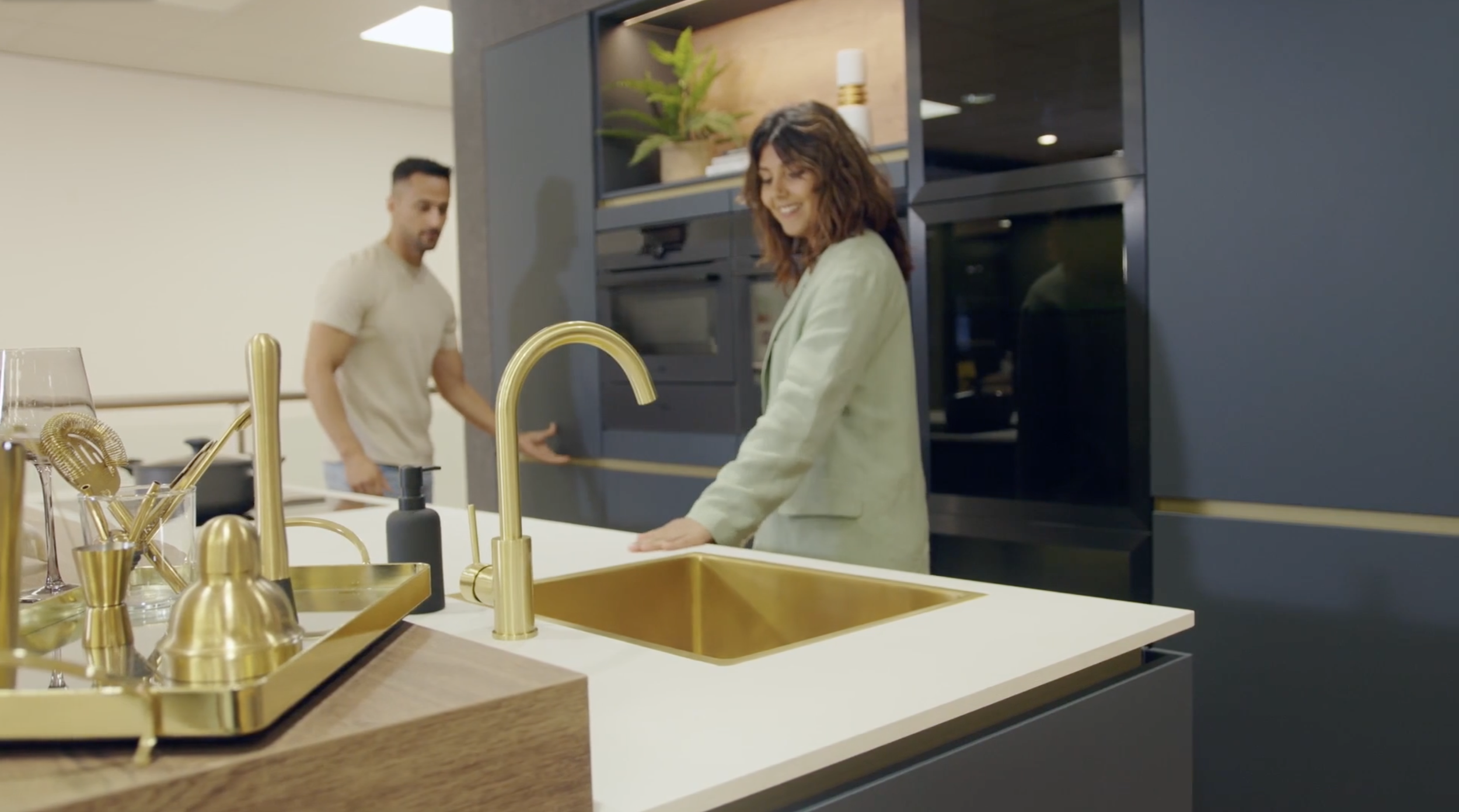 A woman and a man standing in a modern kitchen, with the woman smiling and the man in the background. The kitchen features gold fixtures, a white countertop, and dark cabinetry.