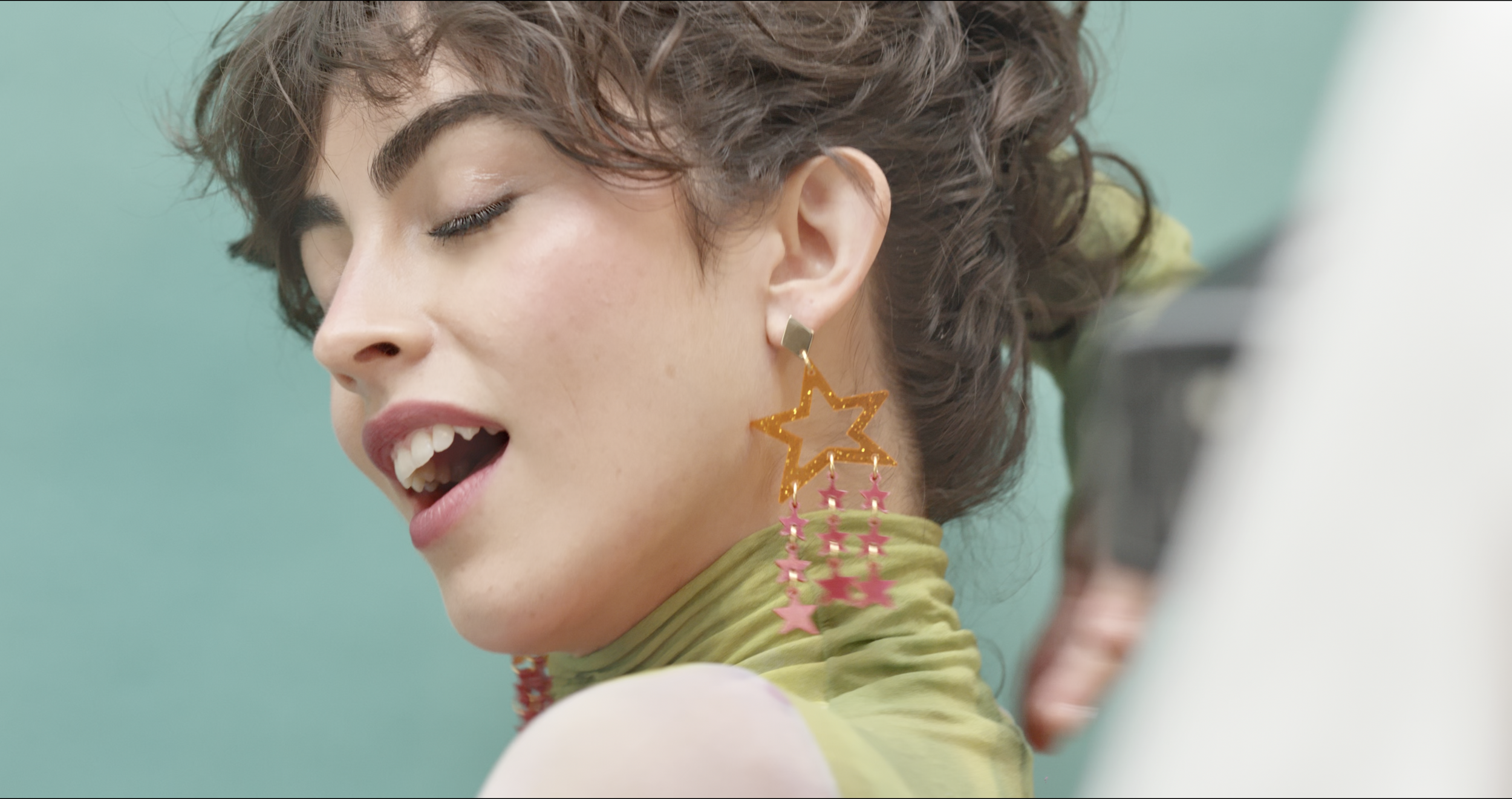 A woman with curly hair wearing large star-shaped earrings with pink star hangings and a yellow top with green accents, posing with her eyes closed against a teal background.