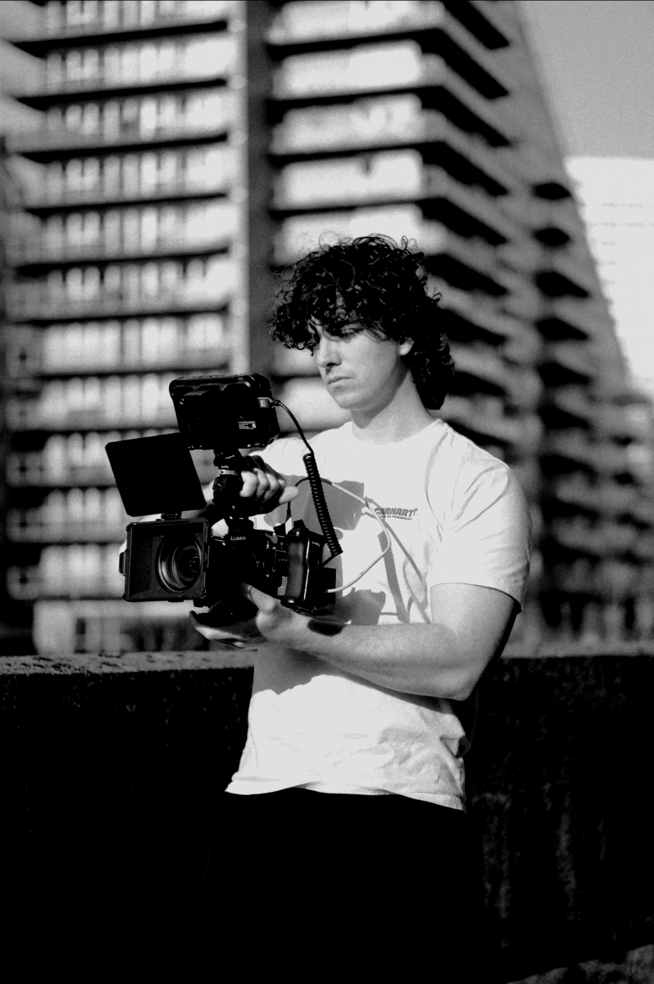 Greyscale image of Luke McCulloch holding a camera with buildings in Manchester behind him