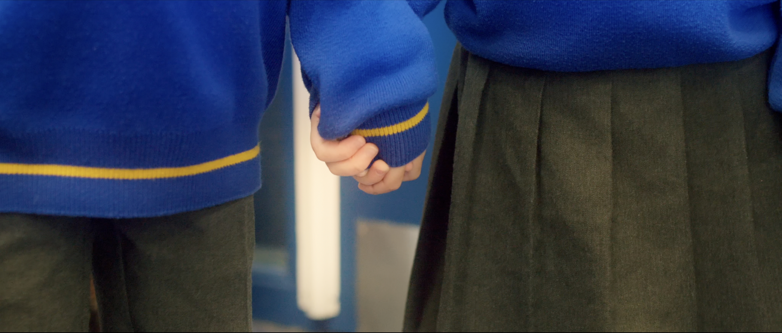 Two children holding hands, wearing blue sweaters with yellow stripes and dark skirts/pants.